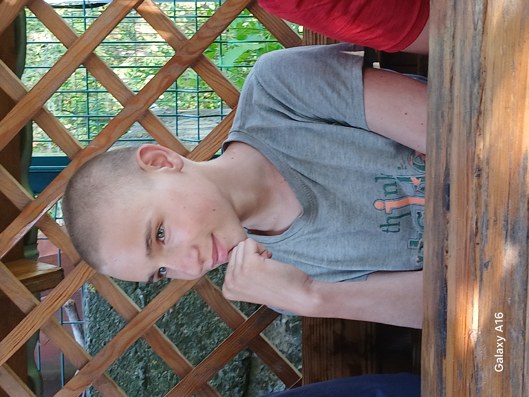 The image shows a close-up of a young person sitting at a wooden table, with a blurred background of a lattice structure and greenery. The person has short, light brown hair and is looking to the left with a slight smile. Their right hand is resting on their chin. They are wearing a gray t-shirt with a graphic design. The setting appears to be outdoors, possibly in a park or garden area. The bottom left corner of the image displays the text "Galaxy A16". The exact location in Ustyvytsia, Ukraine, is not discernible from the image alone.