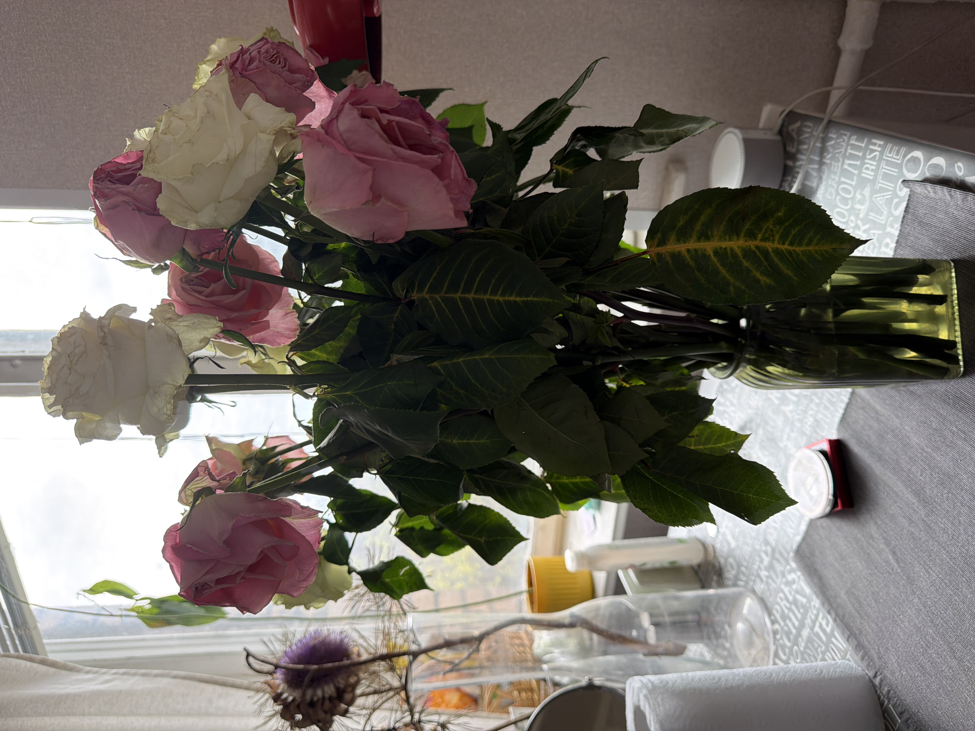 The image shows a vase of pink and white roses sitting on a table near a window. Outside the window, blurry details suggest a generic cityscape. A roll of paper towels, a bottle, and other miscellaneous items are also on the table. The table has a grey tablecloth with a repeating pattern of coffee-related words. There's insufficient detail to definitively pinpoint the exact location in Moscow, Russia. The scene appears to be indoors, likely a domestic setting.