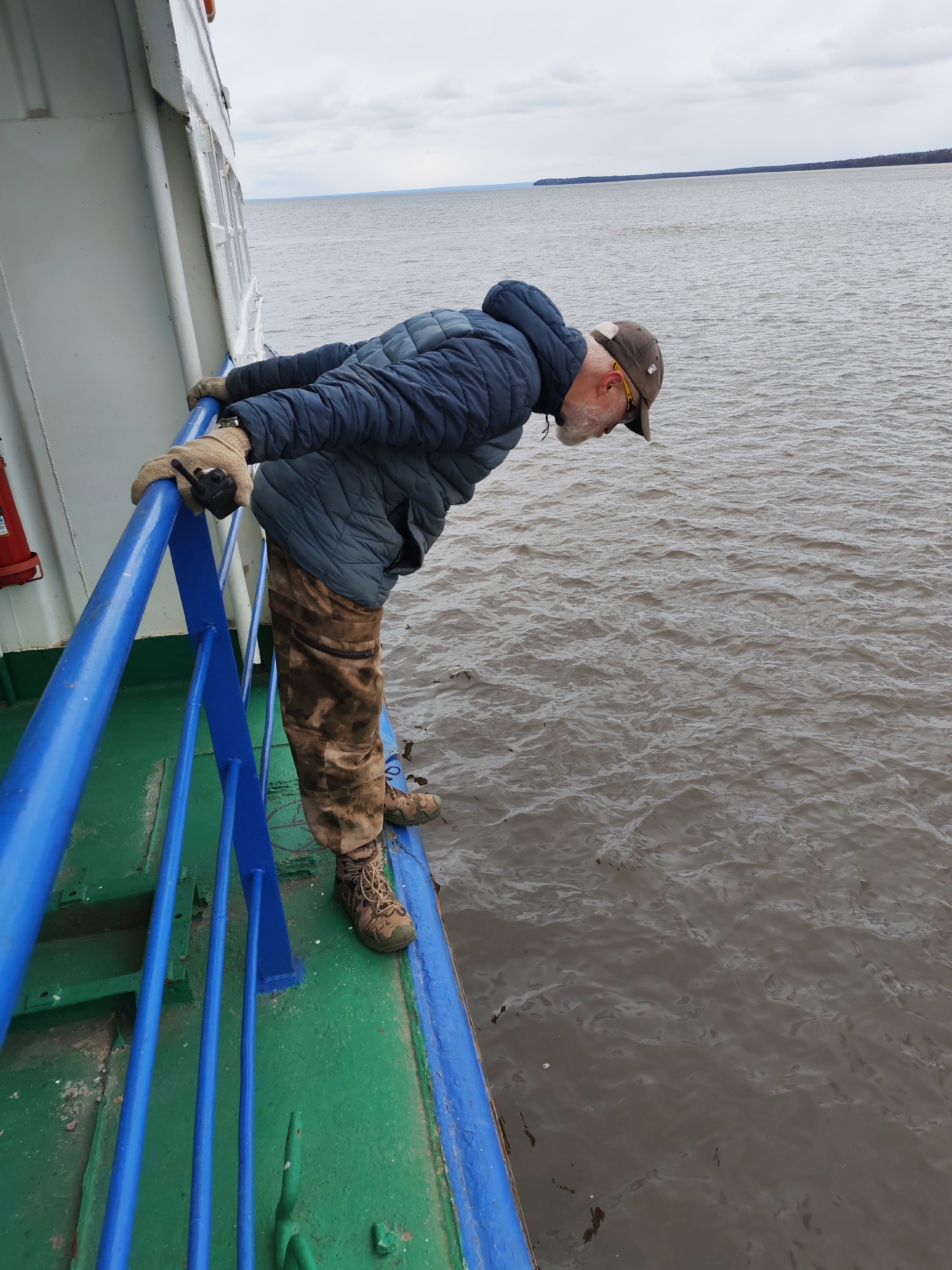 A man with a white beard, wearing a dark blue puffer jacket, camouflage trousers, a brown baseball cap, and light-colored gloves, leans over the blue railing of a boat, looking intently down at the water. A walkie-talkie is clipped to his right glove. The boat's deck is green, and its side is white, with a red fire extinguisher visible on the left. The scene is set on a large body of water, likely a river or lake, with a dark line of trees or land visible on the distant horizon under an overcast, cloudy sky. The water is dark and choppy with ripples, and small pieces of debris are visible floating on the surface. The overall atmosphere is muted, consistent with a cloudy day. The man's posture suggests he is observing something specific in the water below. The location context is Shcheleiki, Russia.