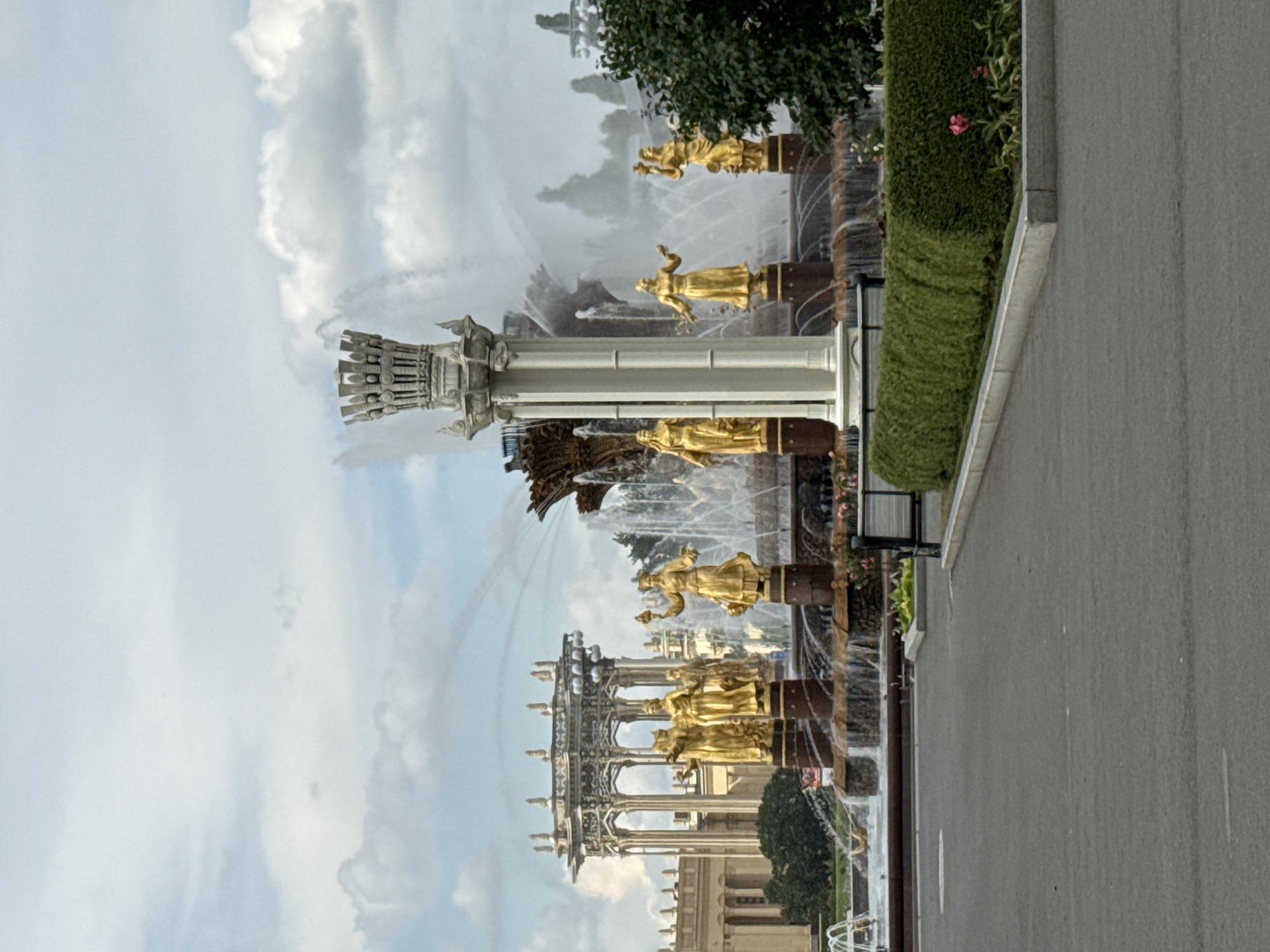 This media file captures a detailed view of a grand fountain with golden statues, likely part of a larger park or architectural complex in Moscow, Russia. The fountain is the central subject, with multiple tiers and ornate golden figures depicting people in flowing robes, holding various objects, and in dynamic poses. These statues are mounted on dark, columnar bases. Water cascades and sprays from various points within the fountain, creating a lively and dynamic scene.
In the foreground, a paved walkway runs alongside a meticulously manicured green hedge and a glimpse of a dark-colored bench. The hedge is lush and features some small pink flowers. Beyond the fountain, hints of other architectural structures and greenery can be seen, suggesting a well-developed public space. The sky above is a mixture of bright white clouds against a pale blue backdrop, indicating a daytime scene with fair weather. The overall impression is one of opulence and public artistry, characteristic of monumental fountains often found in Russian parks and squares. There is no visible text in the image.