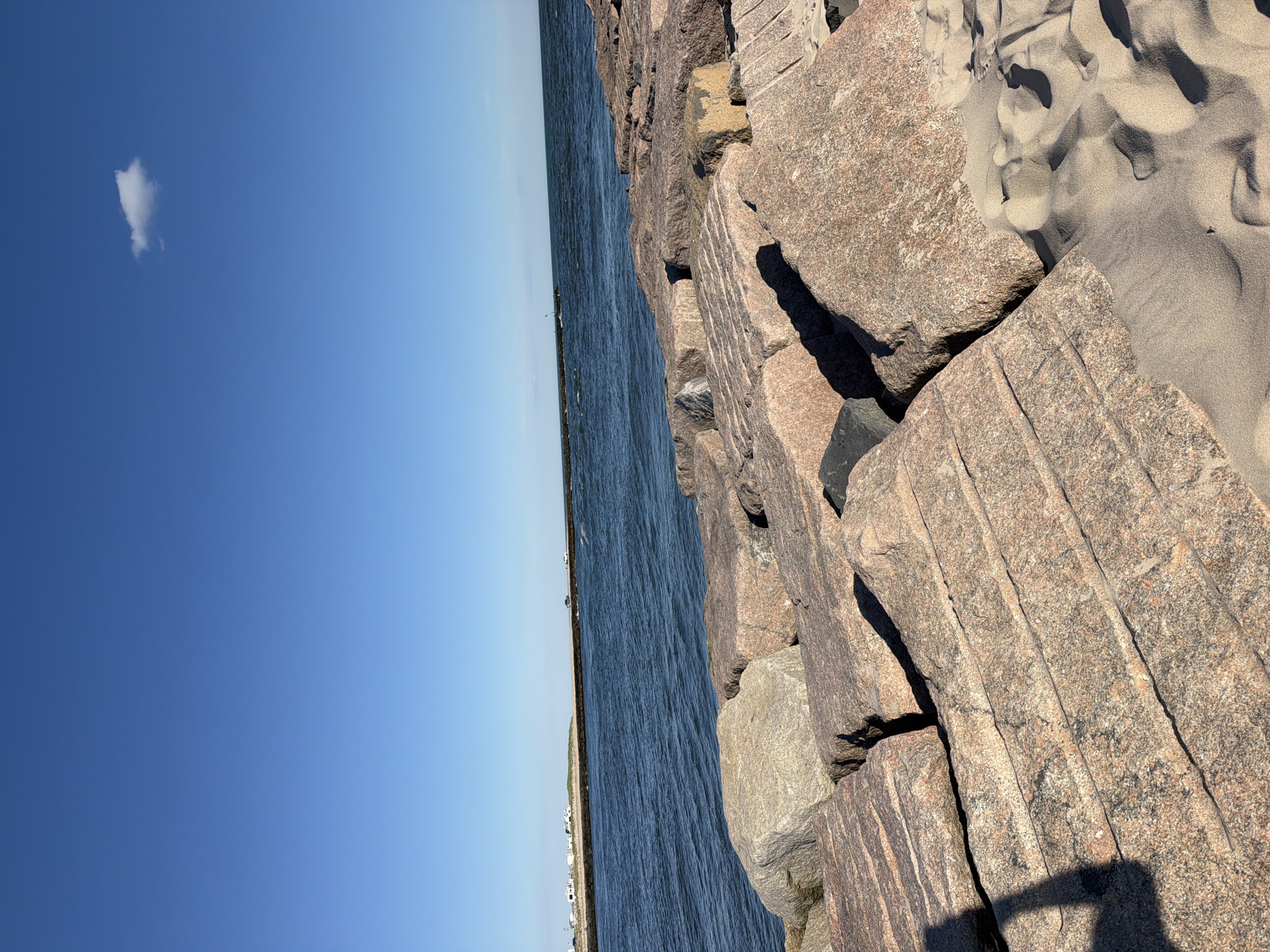 The image shows a rocky shoreline with large granite boulders in the foreground. The ocean is calm and blue, with a distant landmass visible on the horizon. The sky is clear and bright blue, with a single white cloud. The scene appears to be a public beach or park area. There is a shadow of a person on the left side of the frame, suggesting a visitor to the location. The environment is natural and serene, with a sense of open space and a clear view of the water and sky. The location is identified as Hampton Bays, United States of America.