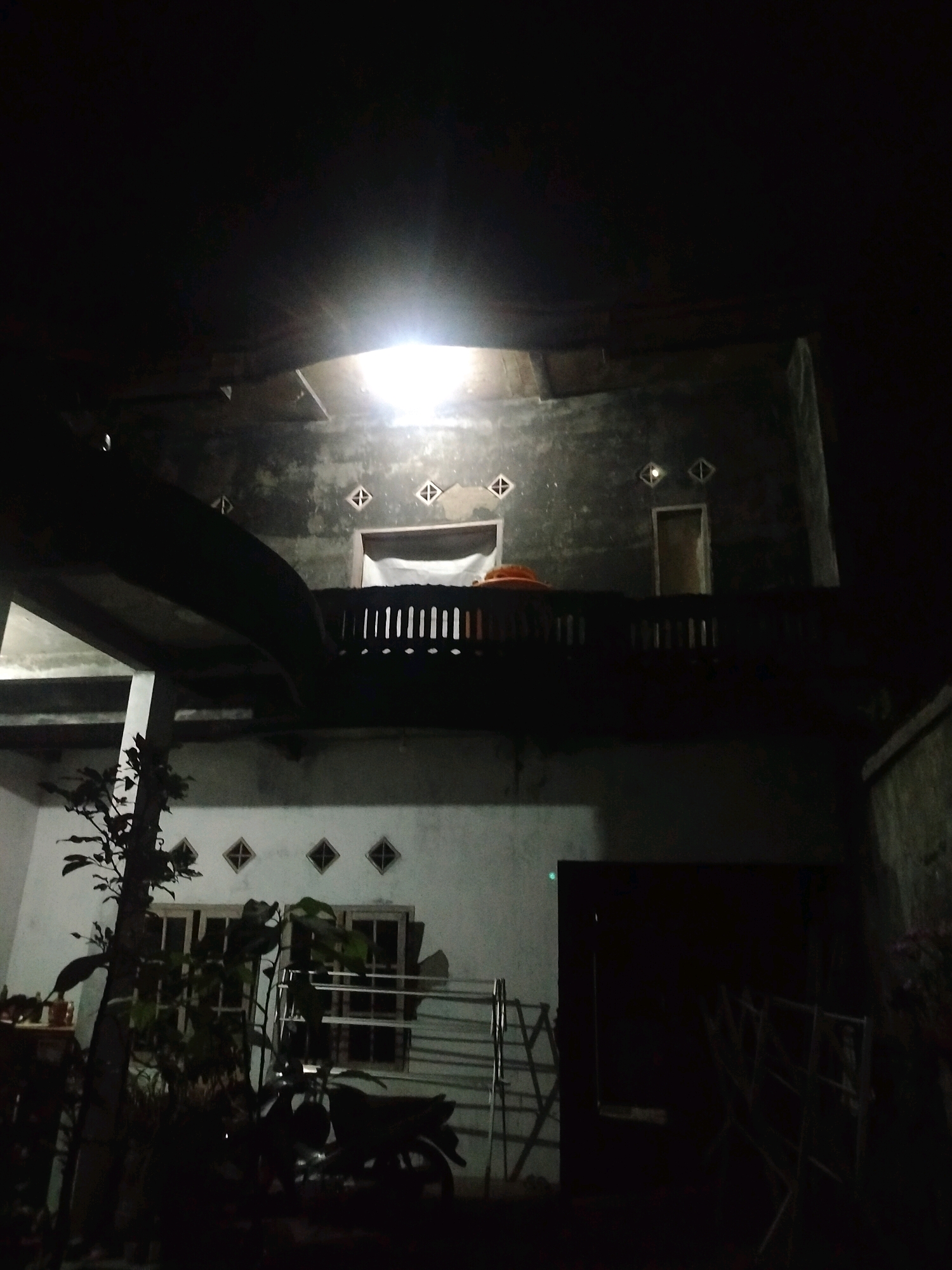 The image depicts the exterior of a two-story building at night, likely in the city of Kroya, Indonesia. A bright light emanates from an overhead fixture on the upper level, illuminating a portion of the building's facade and casting shadows. The upper floor features a balcony with a railing and several diamond-shaped vents. A doorway is visible on the upper level. The lower level of the building has windows with a lattice pattern. In the foreground, a small tree with leaves is present, and a motorcycle is partially visible in the shadows. Metal racks or structures are also present in the scene. The overall environment is dark, suggesting nighttime.