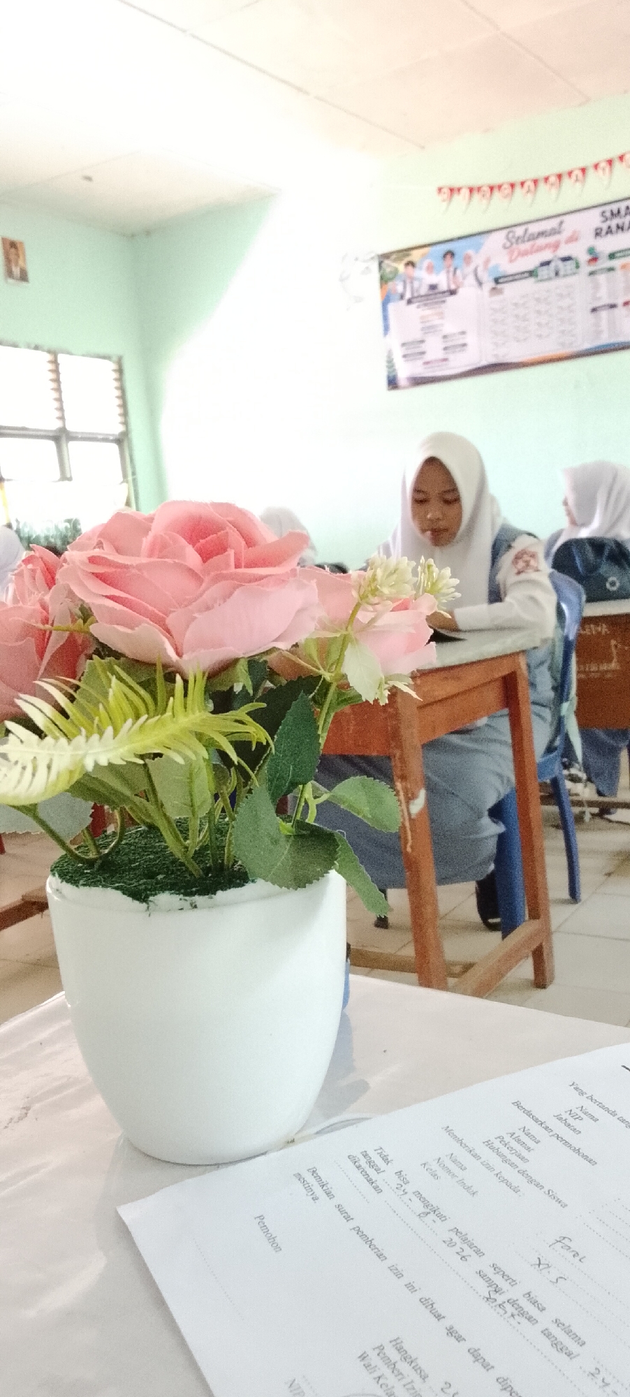 The image captures a classroom scene in Jepara, Indonesia, focusing on a table in the foreground with a white ceramic pot holding a pink artificial rose arrangement. To the right of the flower arrangement, a piece of paper with handwritten text is visible, suggesting a formal document or application. In the background, two young female students wearing hijabs and school uniforms are seated at desks. One student in the middle ground is looking down at her desk, possibly studying or writing. The classroom environment is suggested by the desks, chairs, and posters on the wall, including one with the text "SMA" and "SELAMAT DATANG RA..." indicating it is a school setting. The lighting appears to be natural, coming from the left side of the frame.