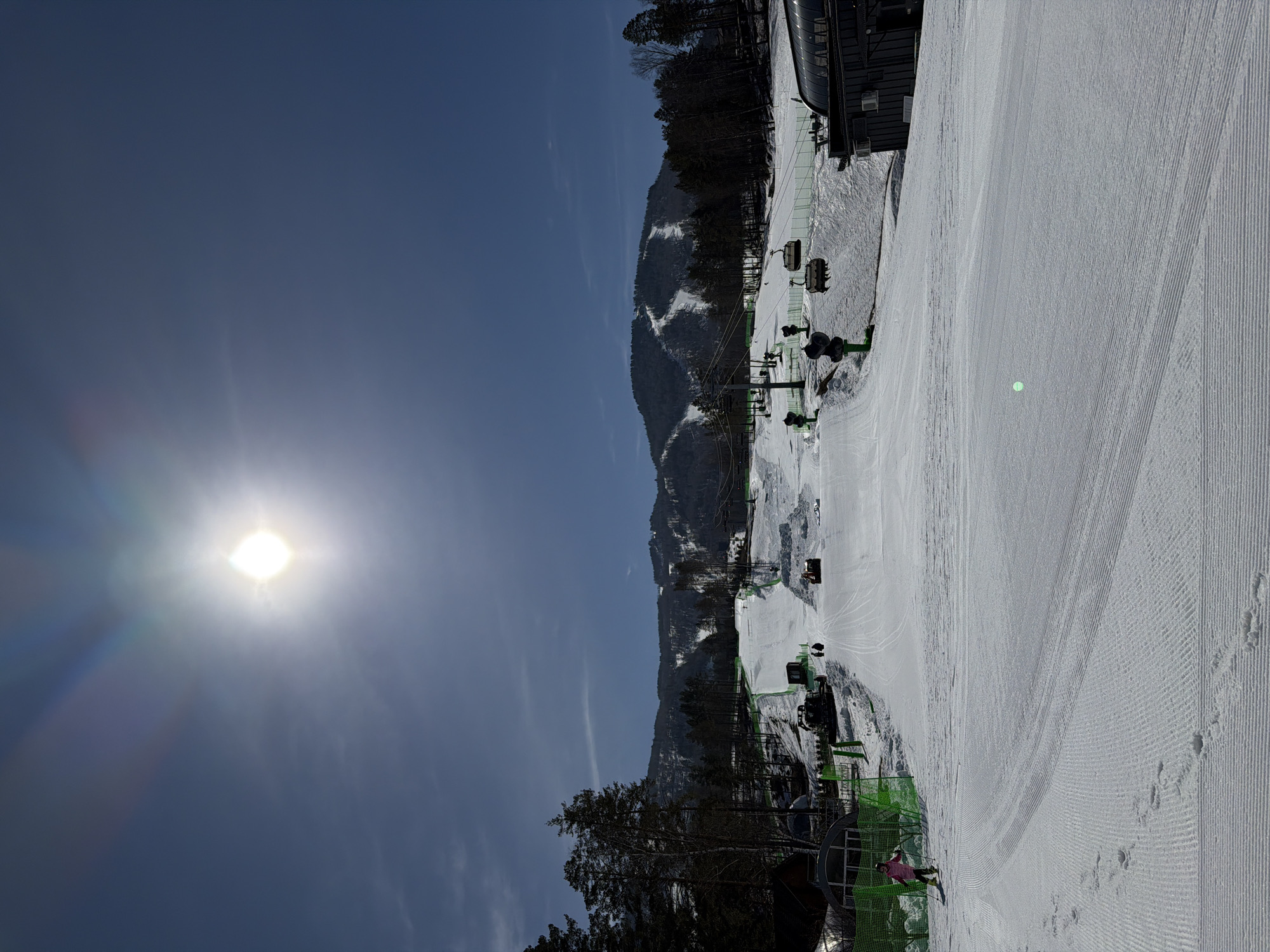 The image depicts a ski slope at a resort in a mountainous region, likely in Озёрное, Russia. The scene is bathed in bright sunlight, with the sun visible in the upper-left corner. The ski slope is well-groomed with clear corduroy patterns on the snow. A chairlift runs along the side of the slope, with empty chairs visible. Green netting lines the edge of the slope. A lone skier in a pink jacket is near the bottom of the slope. The background shows a mountain covered in trees and snow, with visible ski runs.