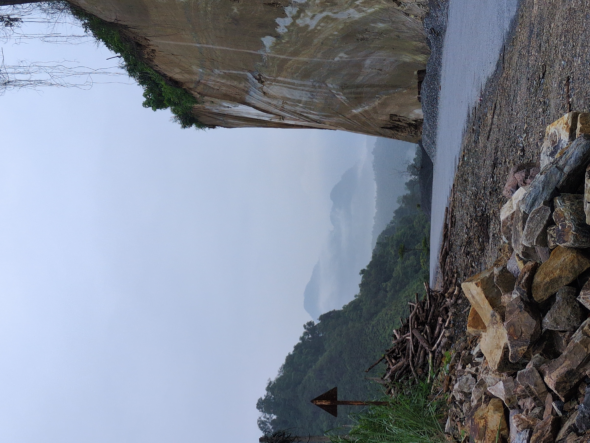 The image depicts a scene in Thuận Hòa Commune, Vietnam, characterized by a dramatic landscape featuring a steep, layered rock face on the right, juxtaposed with lush green hills and misty mountains in the background. In the foreground, a pile of rough-hewn logs and scattered rocks lie on a gravelly surface next to a paved road. A weathered, triangular warning sign stands near the pile of logs. The sky is overcast, contributing to a soft, diffused light across the scene. The environment appears natural and somewhat rugged, with evidence of human activity in the form of the road and discarded materials. There are no people visible in the image.