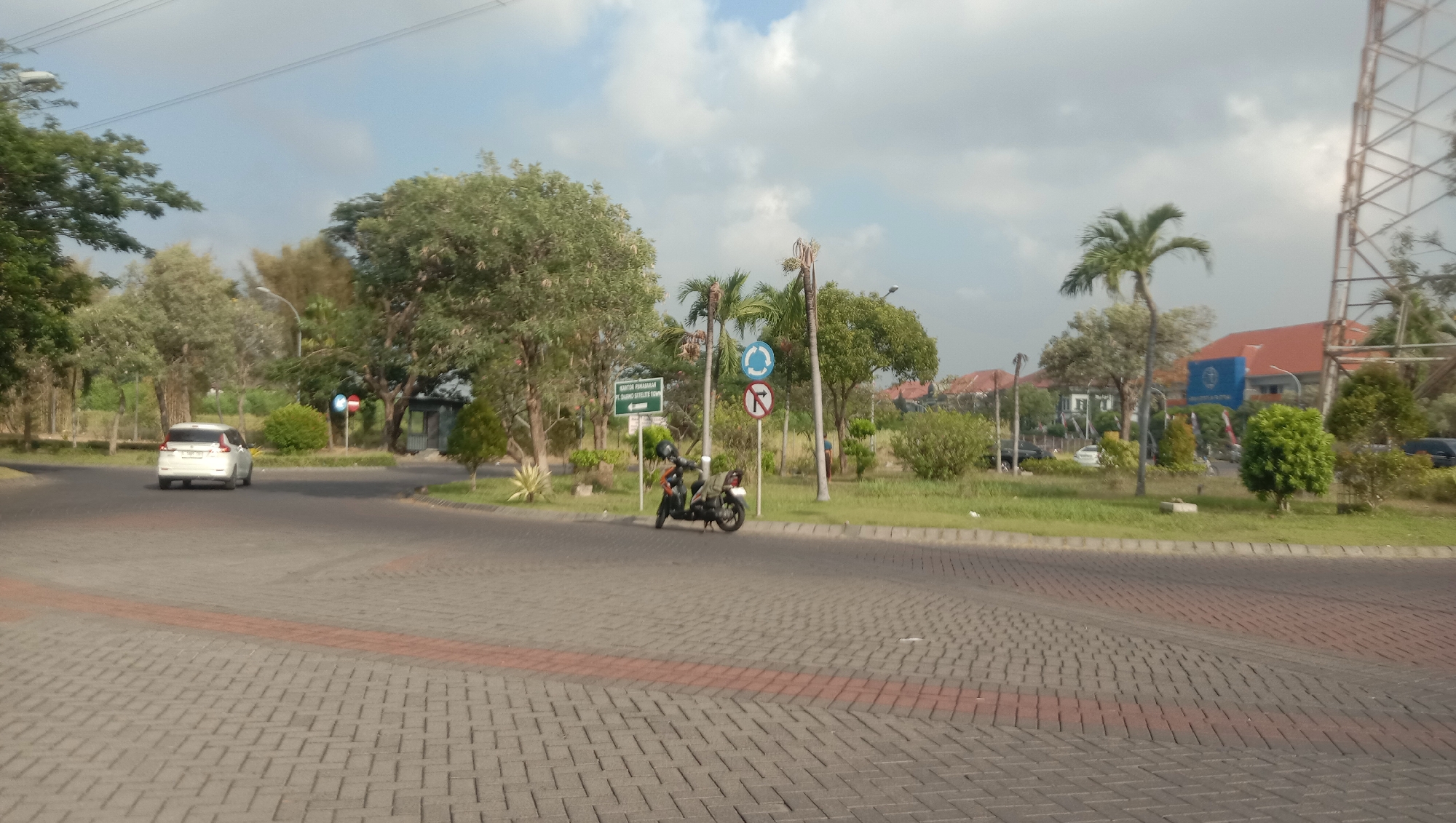 The image shows a paved road with a white car turning left and a motorcycle parked on the side. The road is bordered by a grassy area with trees and palm trees. There are several traffic signs visible, including a directional sign, a circular blue sign with an arrow, and a circular sign with a red line through a turn. In the background, there are buildings with red roofs and a tall tower structure. The sky is partly cloudy.