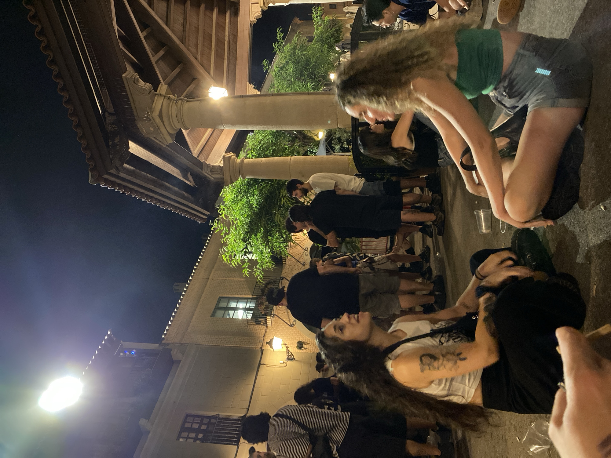 The image depicts an outdoor night scene at the Parc de la Ciutadella in Barcelona, Spain. In the foreground, two young women are seated on the ground, facing each other. The woman on the left has long, dark, curly hair and is wearing a white tank top and black pants, with tattoos visible on her arm. The woman on the right has long, curly, blonde hair and is wearing a green bandeau top and grey denim shorts. Several other people are visible in the background, some standing and some sitting. They are dressed casually in a mix of t-shirts, tank tops, and shorts. The environment includes a building with balconies and windows to the left, and a covered structure with large pillars and a tiled roof to the right. Lighting comes from overhead lamps and a bright spotlight, illuminating the area. Trees and foliage are present, particularly on the right side of the frame. The ground appears to be paved. The overall atmosphere suggests a social gathering or event taking place outdoors in the evening.
