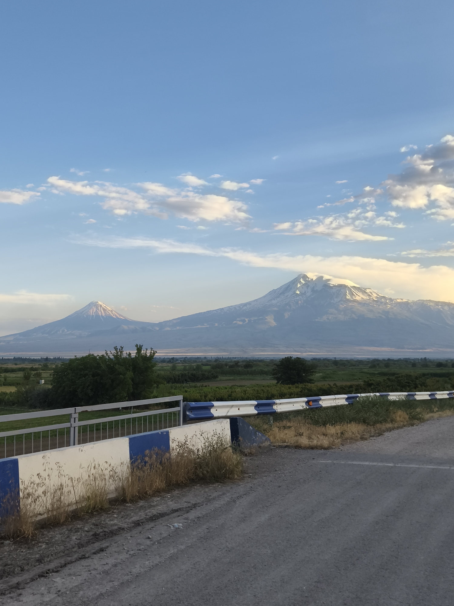 A wide shot captures the majestic twin peaks of Mount Ararat under a clear blue sky with scattered, fluffy white clouds. The larger, right peak is snow-capped, its slopes rendered in soft, muted tones of grey and white, with hints of sunlight catching its summit. The smaller peak to the left is also capped with snow, appearing as a distinct, conical shape against the horizon.
In the foreground, a road with a dirt and gravel surface curves into the scene. To the left of the road, a low barrier made of concrete blocks painted with alternating blue and white stripes separates the road from a field. Dry, tall grass grows along the base of the barrier. Beyond this, green fields and trees stretch towards the mountains, suggesting a rural or agricultural setting. The time of day appears to be late afternoon or early evening, as indicated by the soft, warm light illuminating the mountains and the gentle shadows cast across the landscape. The overall atmosphere is serene and picturesque, highlighting the natural beauty of the location. There is no visible text in the image.