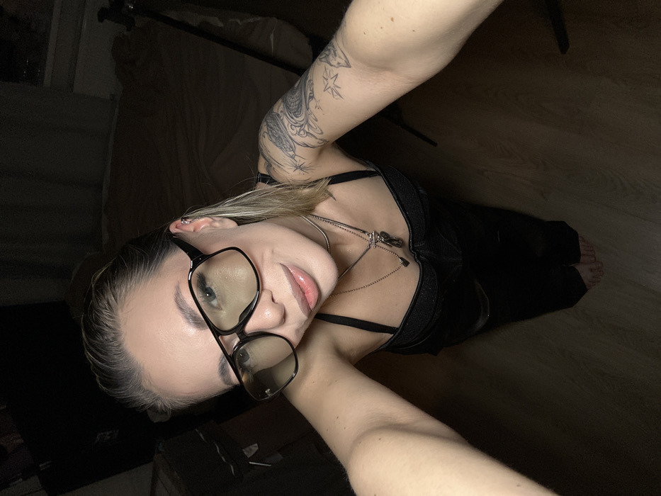 A light-skinned young adult female is depicted indoors in the city of Khimki, Russia. She is taking a high-angle, full-body selfie, looking directly at the camera with a subtle smile and glossy, slightly parted lips. She wears large, dark-rimmed square glasses with light-tinted lenses. Her light-colored, slicked-back hair reveals an ear with multiple piercings. She is dressed in a black textured bustier-style top and black pants, with bare feet visible. Adorning her neck are a thin silver choker and two layered silver-colored necklaces, one with an elongated pendant and another with a toggle clasp and an irregularly shaped dark pendant. Her left arm displays a detailed tattoo sleeve depicting a serpent-like creature, starry elements, and organic patterns. The room is dimly lit, featuring a light-colored wooden floor, a rumpled light-colored fabric surface (possibly bedding), a dark, indistinct outdoor view through a window, and a black tripod in the background.