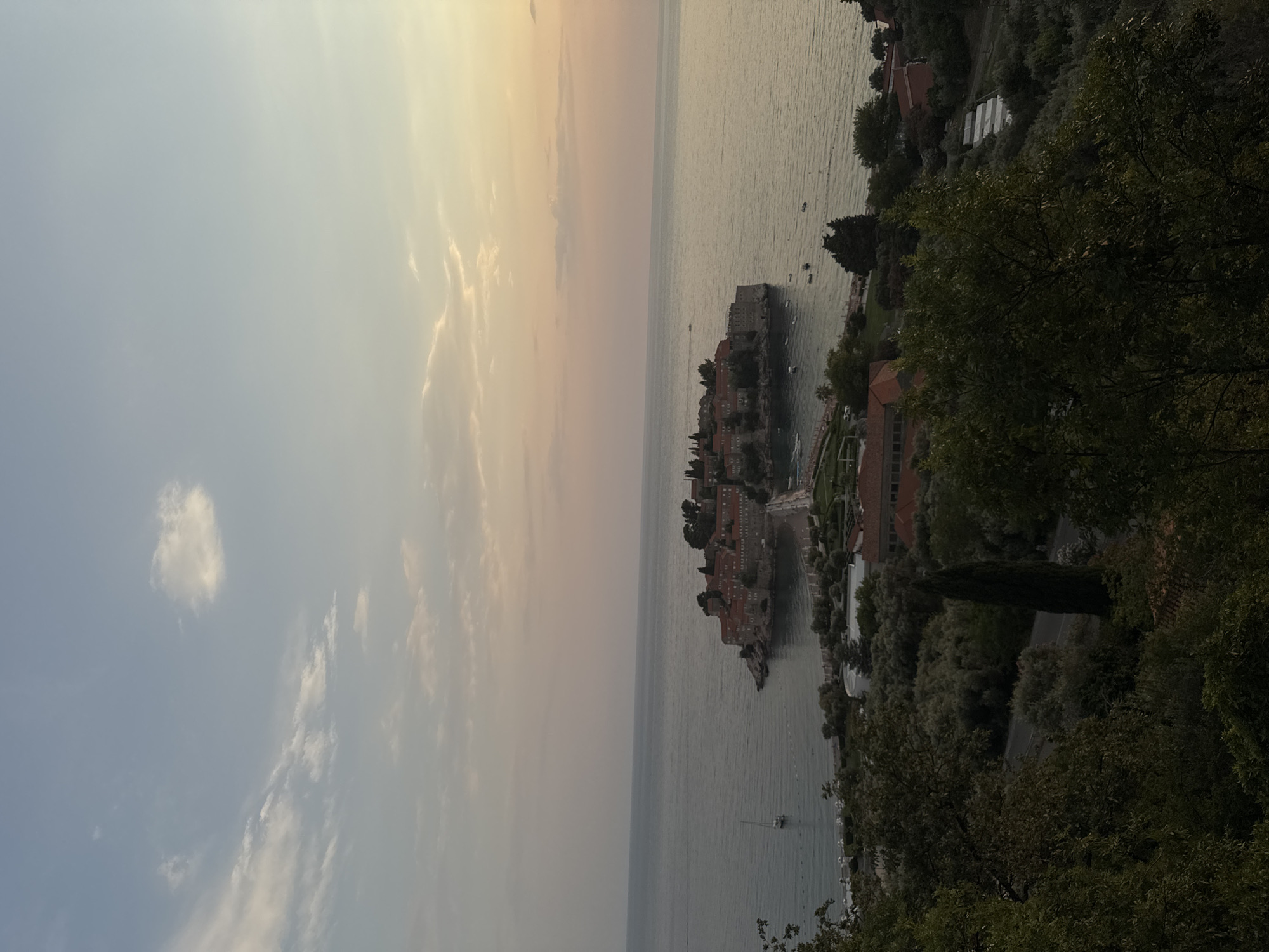 This is a vertical, outdoor photograph of Sveti Stefan, Montenegro, taken during the golden hour of sunset. The image is divided into two halves by the horizon line. The left side of the frame is dominated by the sky, which displays a gradient of soft, pastel colors. Light blue fades into pale yellow and pink hues, with scattered white clouds adding texture and depth.
The right side of the frame features the sea and the iconic Sveti Stefan island, a picturesque islet connected to the mainland by a narrow causeway. The water is calm and reflects the soft light of the sunset, with gentle ripples visible. Several small boats are dotted on the water. The island itself is densely packed with historic stone buildings, many with red-tiled roofs, nestled amongst lush greenery. The causeway is also visible, leading to the island. On the mainland, more buildings and dense trees line the coast, with a dark green forest in the foreground obscuring a portion of the view. The overall atmosphere is serene and tranquil, conveying a sense of natural beauty and historical charm. The time of day is clearly sunset, with the soft, warm light illuminating the sky and sea.