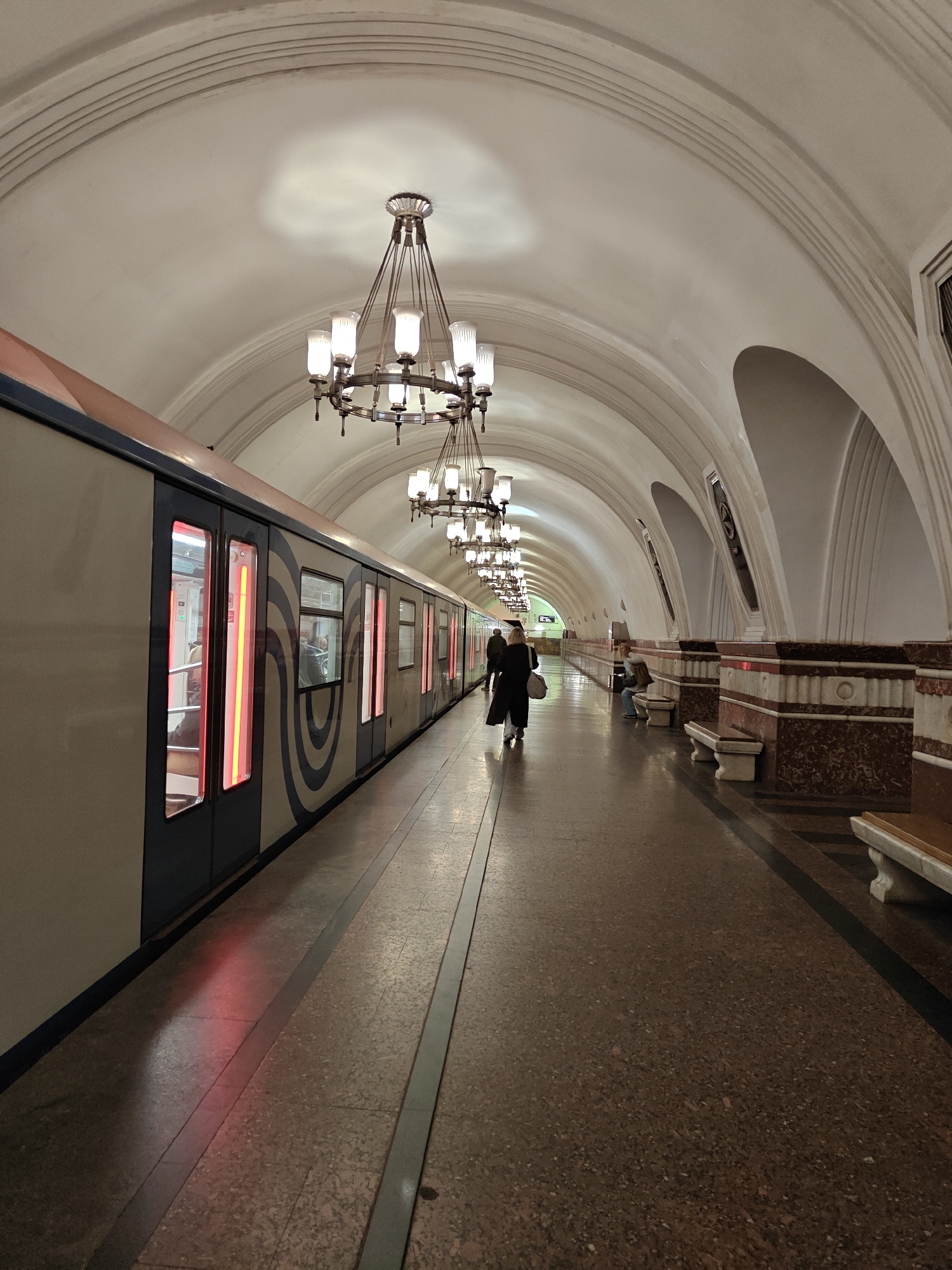 The image depicts a Moscow, Russia metro station platform. A train is stopped on the left. A person walks away from the camera towards the end of the platform. Other people sit on benches along the right wall. Chandeliers hang from the arched ceiling. The platform floor is speckled. The walls are decorated with marble and other ornate details.