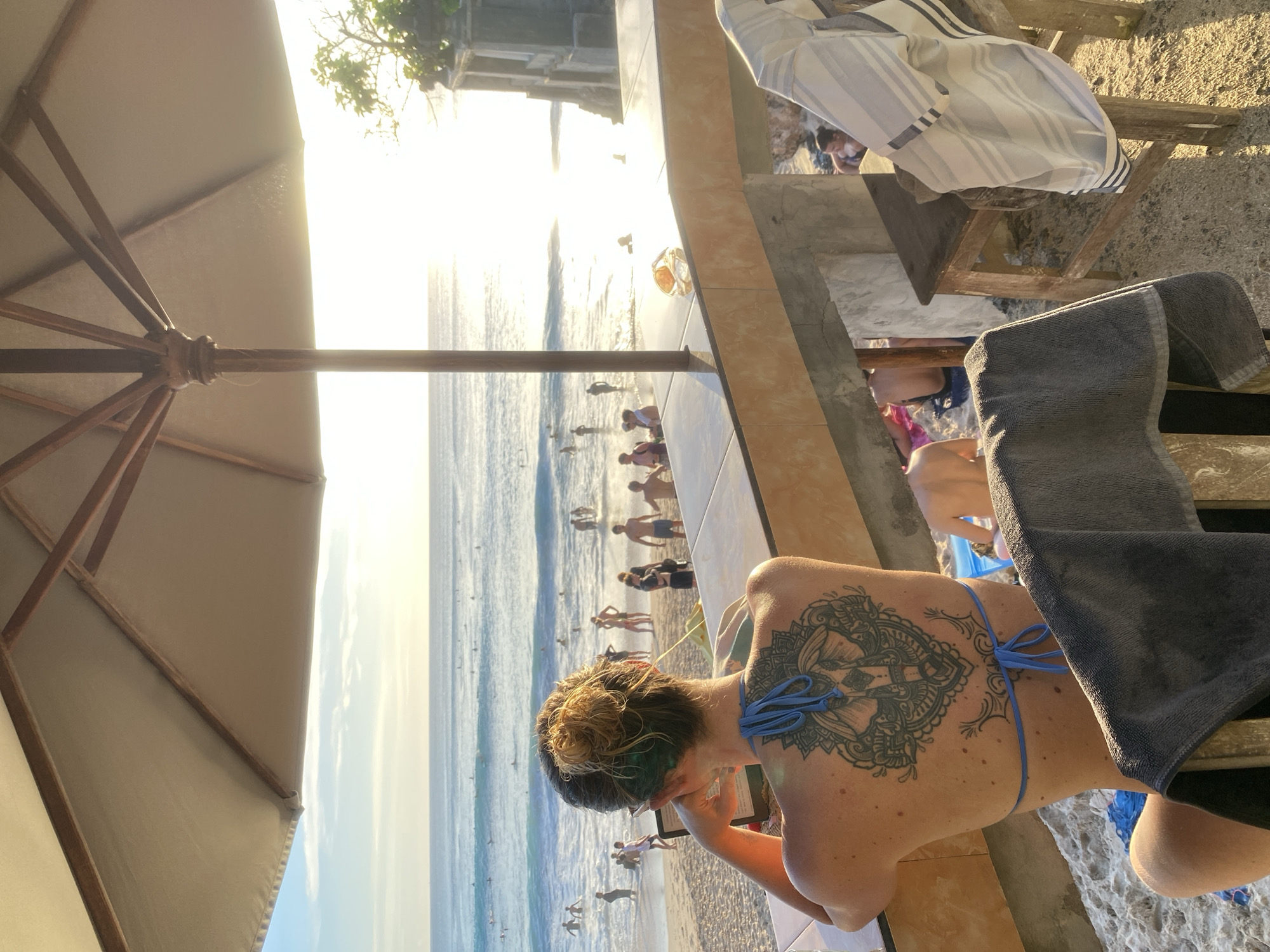 The image captures a vibrant beach scene during the golden hour, likely sunset, from an elevated vantage point. In the immediate foreground, a large beige beach umbrella on the left provides shade, while on the right, a lounge chair draped with a gray towel indicates a relaxing spot.
The primary subject is a woman seen from behind, seated in the lounge chair. She features a prominent, intricate mandala-style tattoo on her upper back and shoulder blades. Her hair is styled in a messy bun, with noticeable green or blue highlights. Dressed in a blue string bikini top, she appears engrossed in a small electronic device, possibly an e-reader or phone, held in her hands. Beside her, on a concrete and tiled ledge, a golden-colored glass, possibly containing a drink, is visible.
Beyond this elevated seating area, a bustling beach unfolds. Numerous people are scattered across the sandy shore and in the ocean, where gentle waves can be seen. Many individuals are swimming, wading, or walking along the water's edge. The low sun, positioned near the horizon, casts a brilliant, warm glow across the water, creating shimmering reflections and bathing the entire scene in a soft, ethereal light. The clear sky meets the ocean in the distance. The setting suggests a popular coastal location, consistent with Kuta Selatan, Indonesia, offering a serene and leisurely atmosphere. No discernible text is visible on any objects or surfaces within the frame.