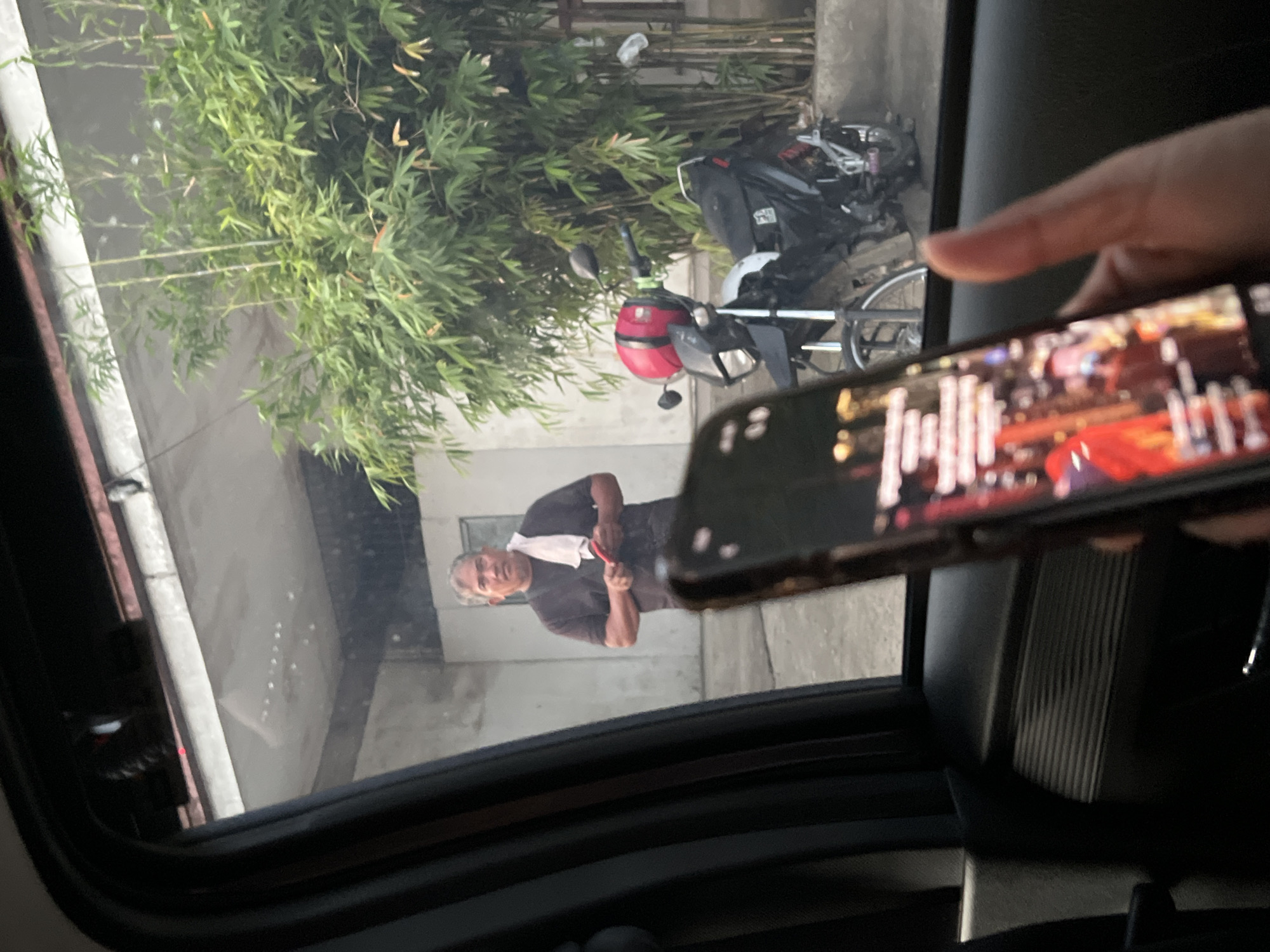 The image, seemingly taken from inside a car, shows an older man standing outside, near a motorcycle. The man is wearing a dark shirt and has a white towel around his neck. He is holding something red in his hand. The background features a building facade and some foliage. A phone is in the foreground, held by a hand, displaying content on its screen. The location is likely Davao City, Philippines.