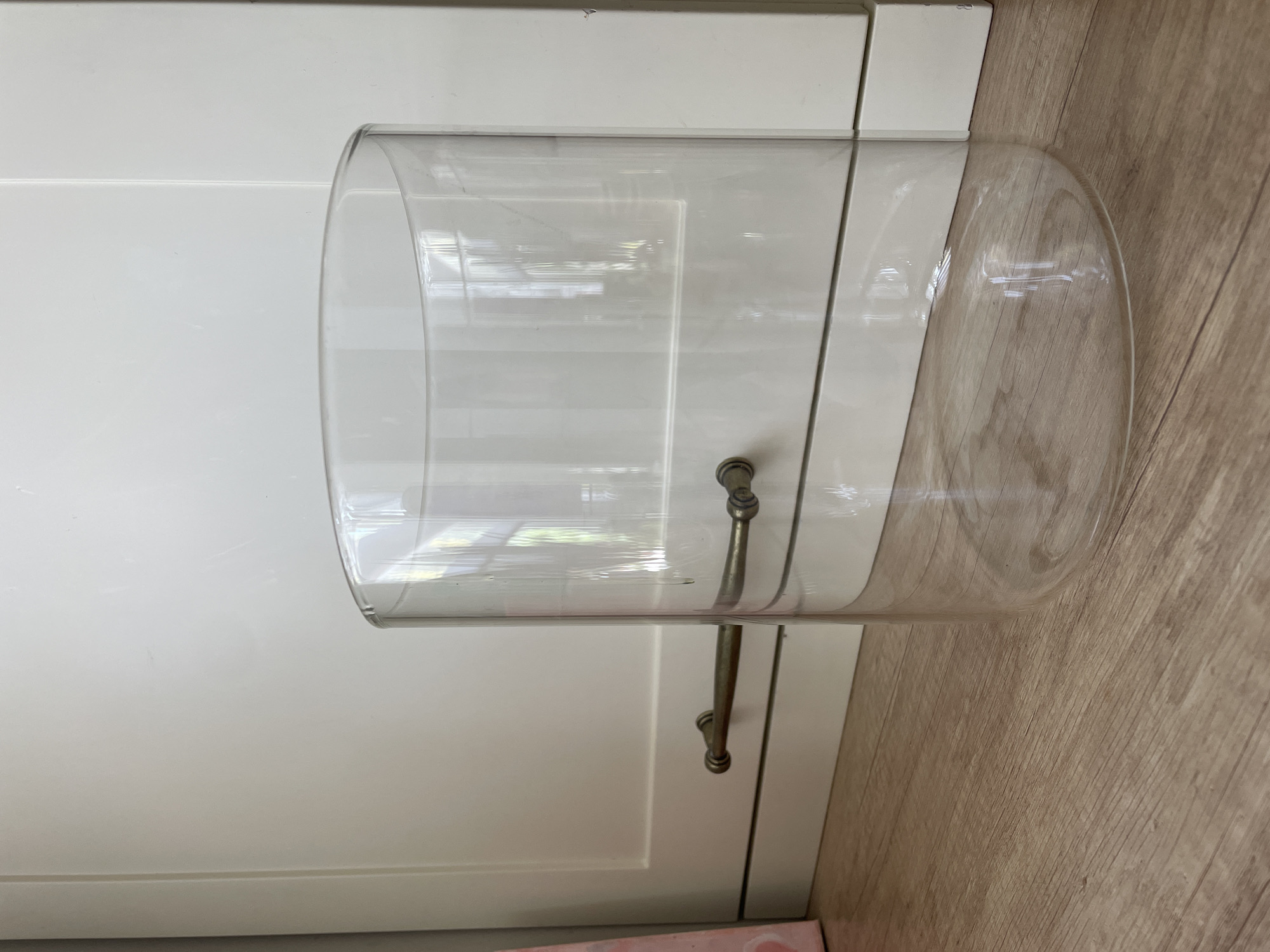 A clear, cylindrical glass container rests on a light-colored wooden floor, positioned horizontally. The container appears to be made of thick glass and has a domed end on the right side. Two metal handles, possibly made of brass or antique silver, are attached to the outside of the glass. The handles have a decorative, knob-like end and are connected to the glass by screws or rivets.
Behind the glass container is a white surface, likely cabinet doors, with visible paneling lines and a subtle sheen that reflects the light. The overall scene is indoors, suggested by the presence of the cabinet and the floor. The lighting appears to be bright, casting reflections on the glass and highlighting the texture of the wooden floor. The image is taken from a slightly elevated angle, looking down at the object. There are no people visible in the image. The environment suggests a domestic setting, possibly a kitchen or living area. There is no visible text in the image.