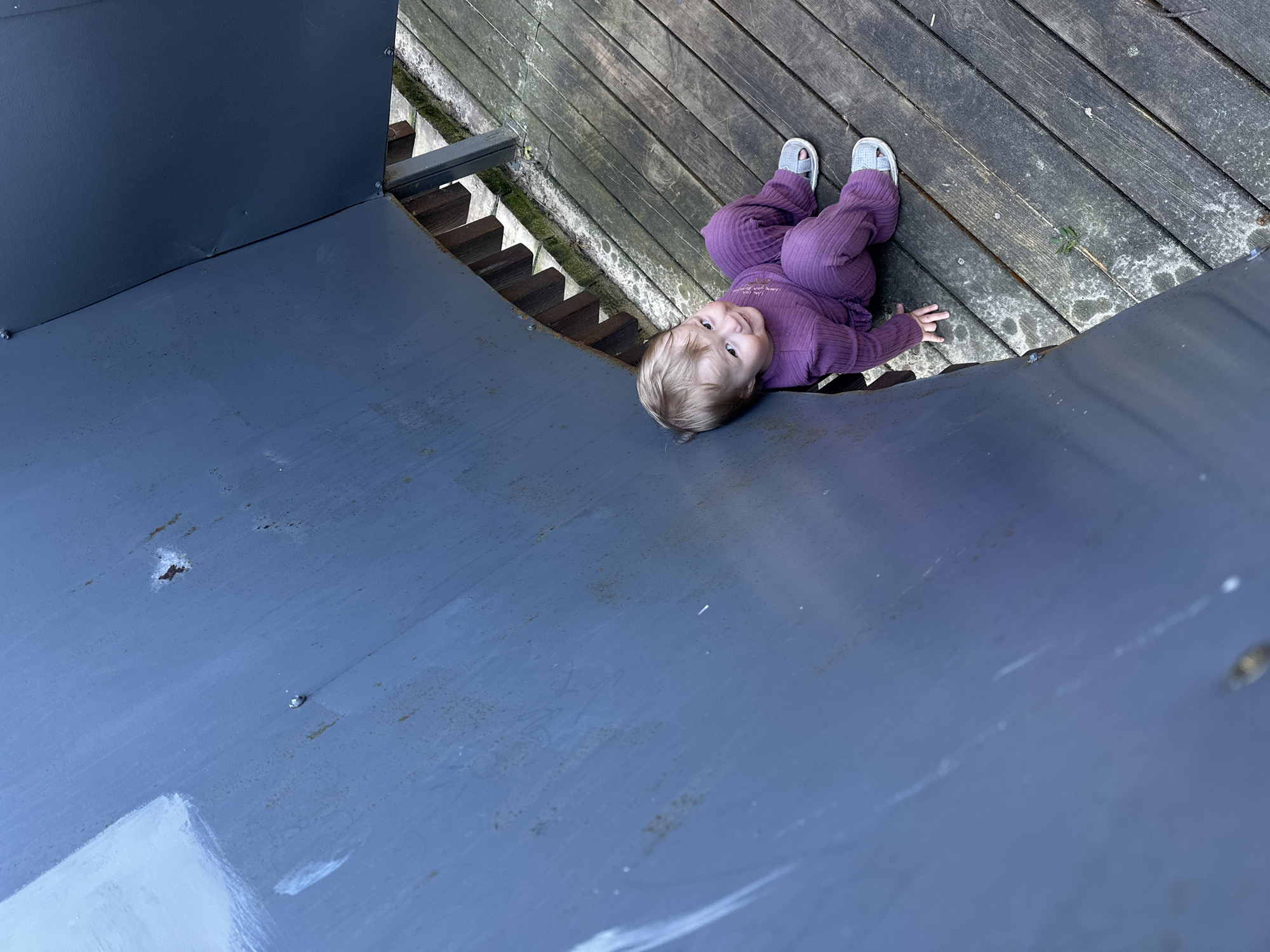 An overhead view captures a young child lying on their back on a weathered wooden deck in an outdoor setting in Nizhny Novgorod, Russia. The child, with light blonde hair, is dressed in a purple ribbed long-sleeved top and matching pants, paired with light gray sandals. They are looking directly up towards the camera with a slight, content smile. Faint white text, which appears to read "I am your sun" followed by a small heart, is visible on the child's top.
The wooden deck consists of dark, aged planks, with some moss and damp patches evident in the crevices. To the left and in the foreground, a large, curved surface painted a dull blue-gray color dominates the frame. This surface, possibly metal or painted concrete, shows minor scuffs, dirt, and some small rust spots. Metal structures or railings are visible where this blue-gray surface meets the wooden deck. The image is taken from an elevated perspective, suggesting the child is on a platform or a play structure. The scene is illuminated by natural light, indicating it is daytime.