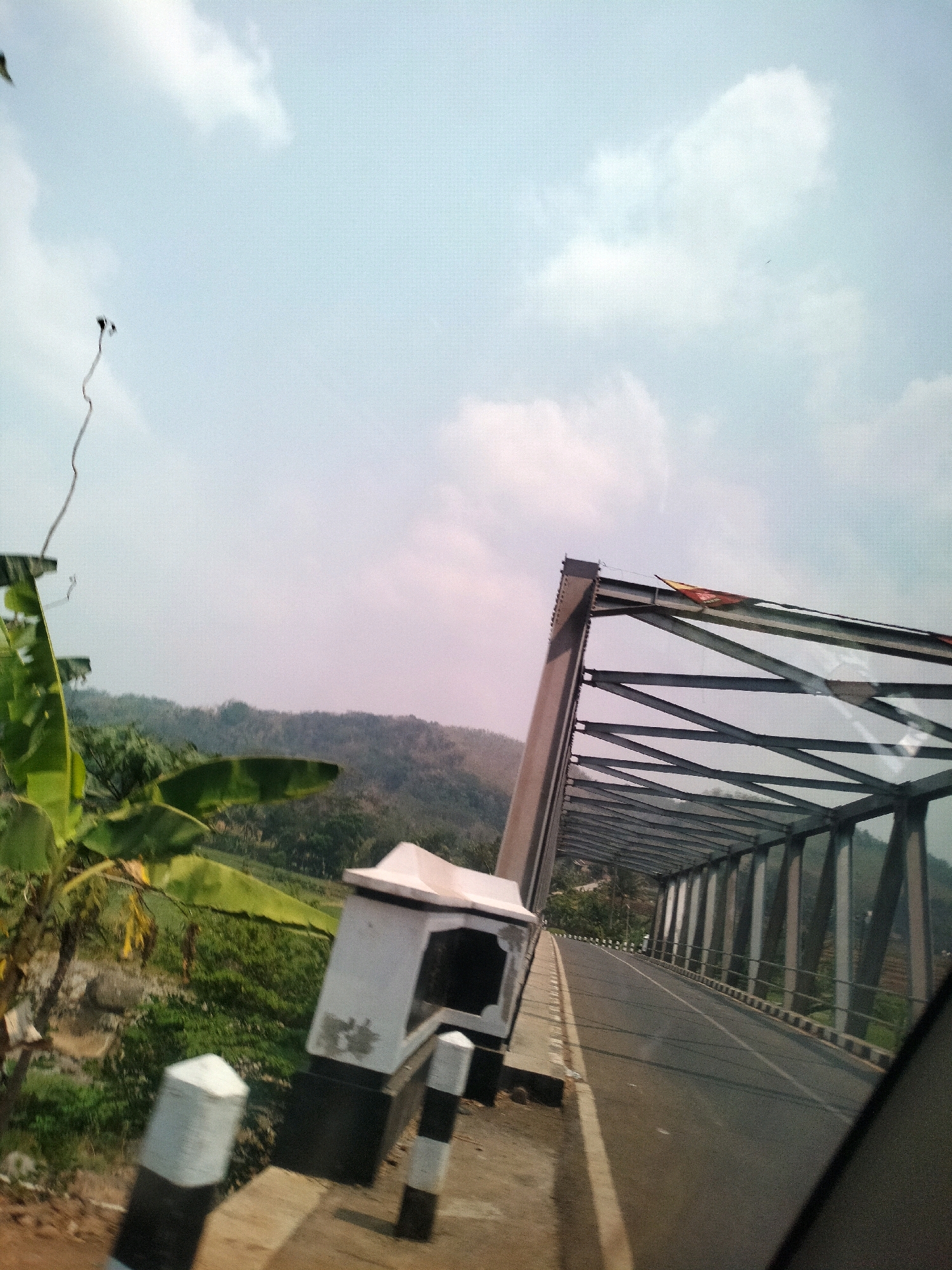 The image is a medium shot, taken from a moving vehicle, showing a steel truss bridge in Madugowongjati, Indonesia. The bridge spans across a rural landscape of rolling hills covered in trees. A two-lane asphalt road leads onto the bridge. On the left side of the road, there are some banana trees and lush green vegetation. A small, white structure with black stripes, possibly a traffic control booth or signage post, is visible next to the road. The bridge itself is constructed with dark grey steel beams and supports. A few colorful flags are attached to the top of the bridge structure. The sky is a pale blue with scattered white clouds. There are no people clearly visible in the image. The overall scene depicts a scenic view of a bridge in a natural, rural environment.