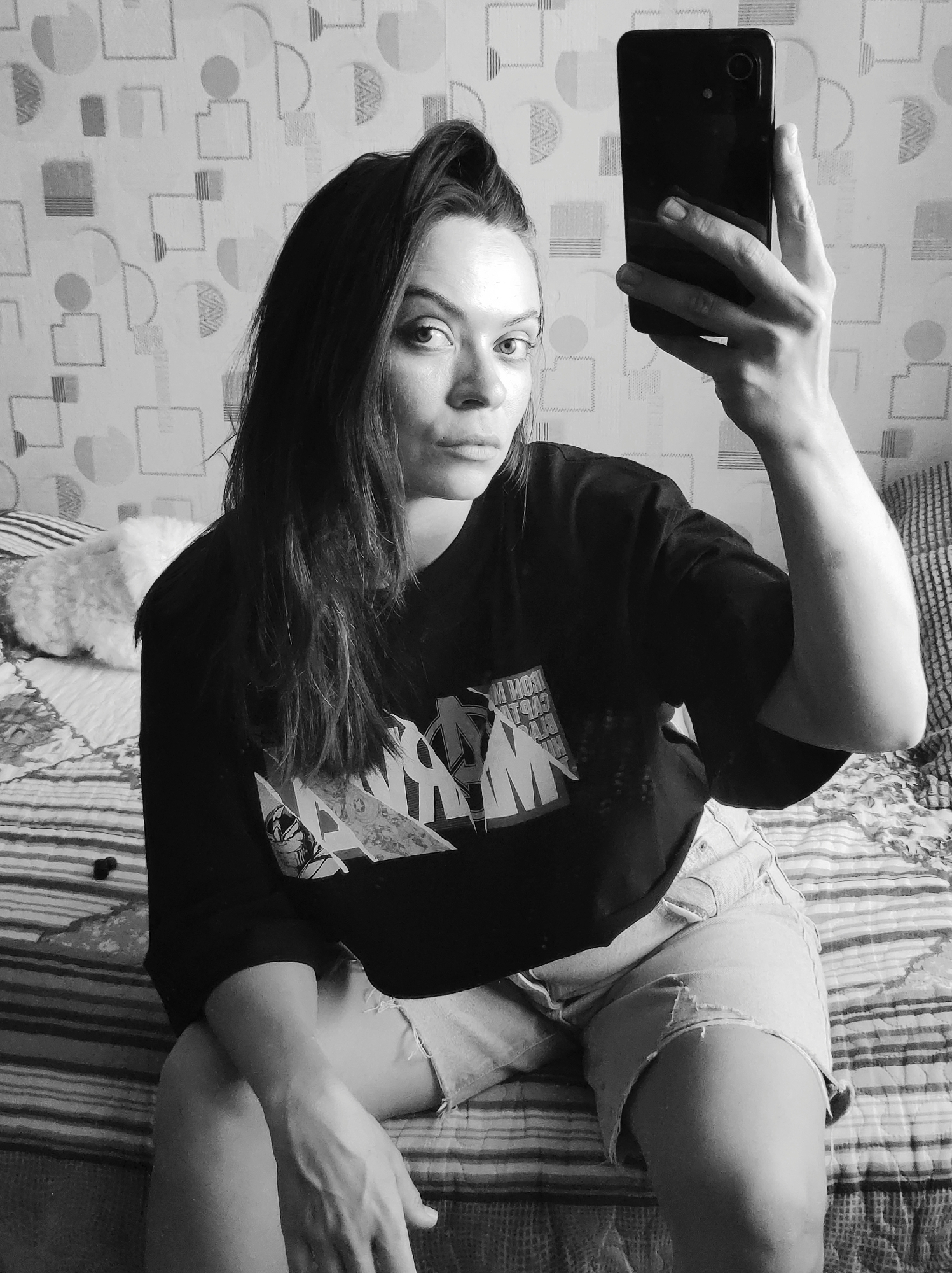 A medium shot photograph in black and white shows a young woman seated on a bed. She is holding a smartphone up and taking a selfie. She is wearing a dark T-shirt with a graphic design on it and light-colored denim shorts. Her hair is long, dark, and parted to the side with a slight lift at the front. The room includes patterned wallpaper and a striped bedspread. Two small, dark items are on the bed. It is not possible to definitively determine that the location is Chaikovsky, Russia from the image.