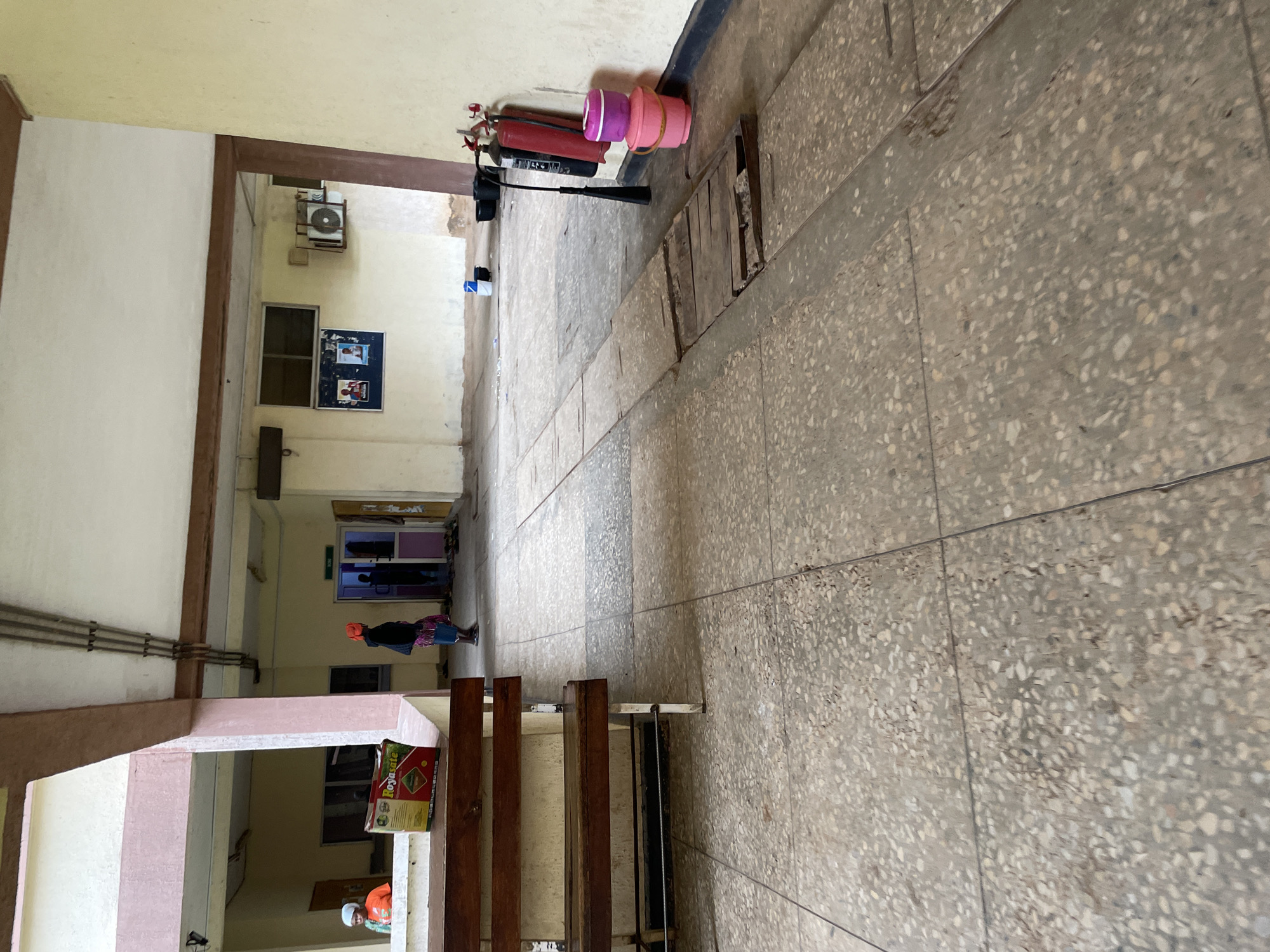 The image depicts a covered outdoor walkway with a terrazzo floor. On the left, a wooden bench is positioned against a wall, with a box of "Royasate" products on it. A person wearing an orange top and white head covering is visible in the background to the left. Further down the walkway, to the right of center, two individuals are seen entering a doorway. One of them wears an orange headscarf. Adjacent to the doorway, on the wall, are two fire extinguishers and a stack of pink buckets. The walkway is lined with concrete pillars and beams supporting the roof. The environment appears to be part of a building complex, possibly in Gwong, Nigeria, characterized by its weathered materials and functional design. The scene is static, with no overt actions or interactions occurring.