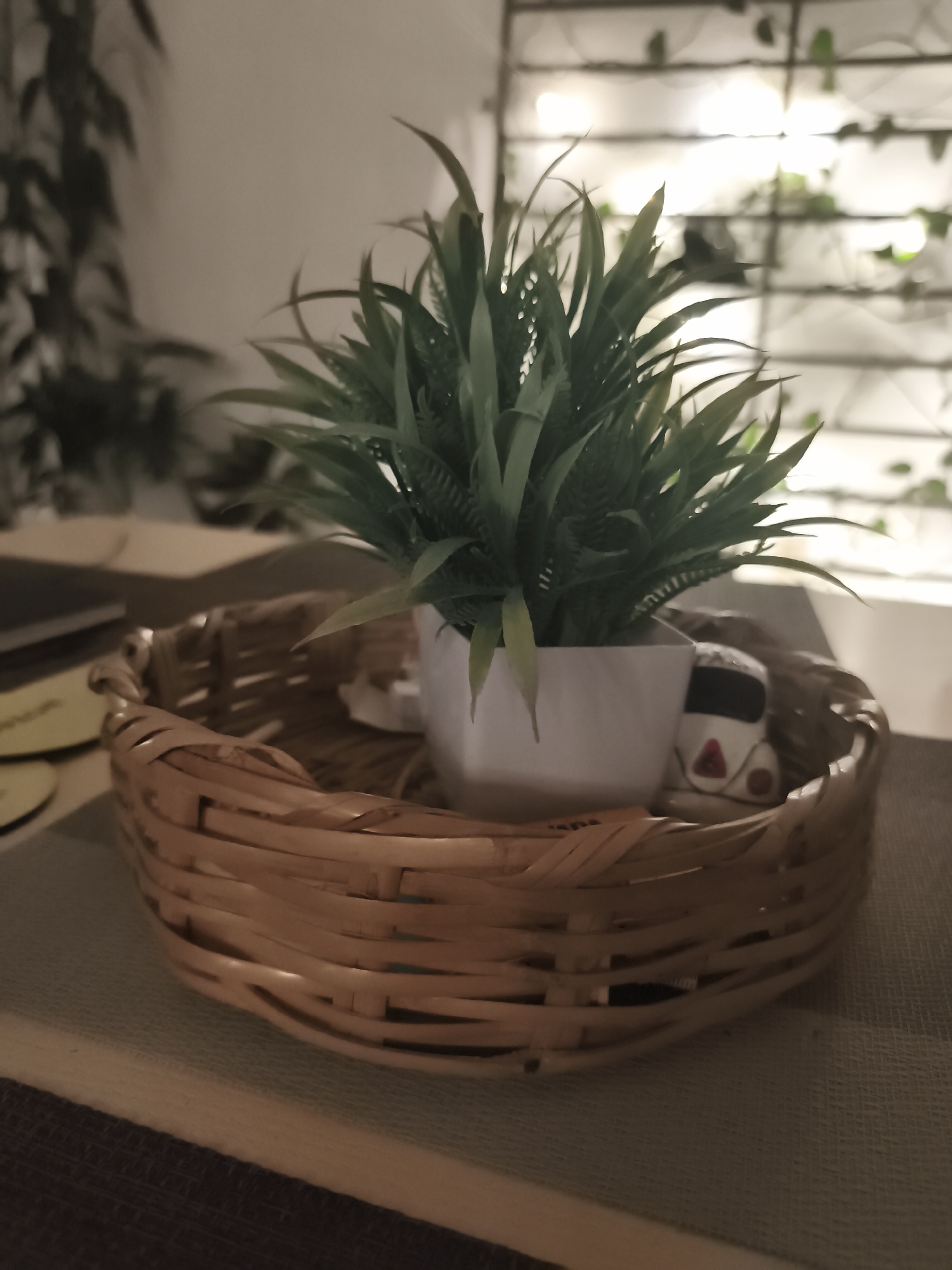 A close-up photograph shows a woven wicker basket on a surface, with a small, square white planter containing an artificial green plant inside the basket. A small decorative white Volkswagen Beetle figurine is positioned near the plant. The background is slightly blurred, showing a wall with a grid pattern of horizontal and vertical lines, and some indistinct foliage. The lighting suggests an indoor setting. No people are visible. The location within Mexico City, Mexico is not evident from the image.