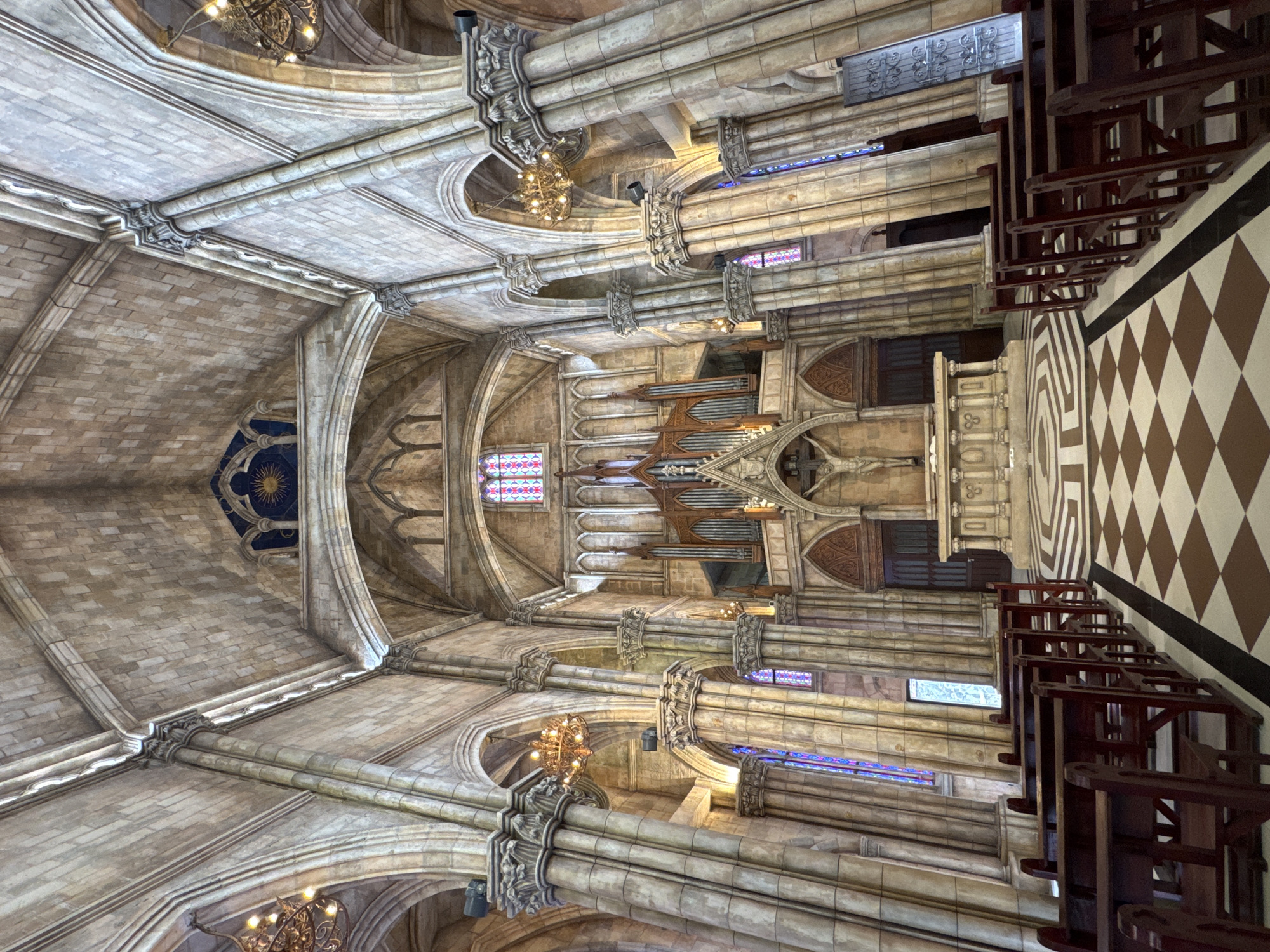 The image depicts the interior of Da Nang Cathedral, a Catholic church in Đà Nẵng, Vietnam. The view is oriented upwards, capturing the architectural details of the vaulted ceiling, adorned with intricate stonework and arches. Chandeliers hang from the ceiling, illuminating the space. Along the sides, columns support the structure, with stained glass windows visible. An organ is positioned above the altar. In the foreground, rows of wooden pews are visible, and the floor features a checkered tile pattern. No people are visible in the frame.