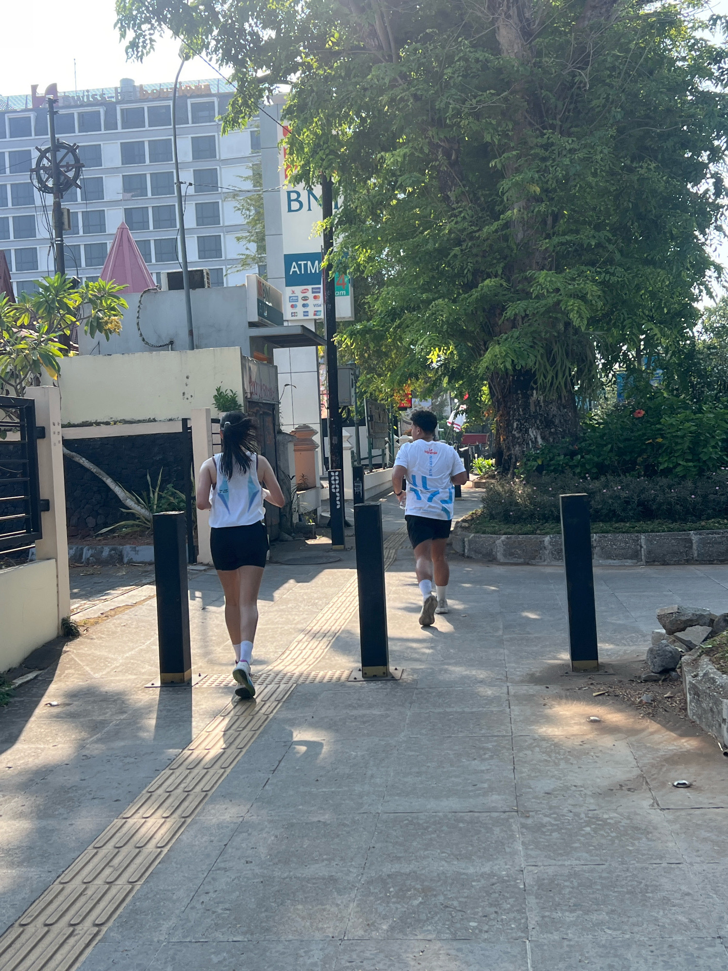 Two individuals are jogging on a paved sidewalk adjacent to a tree-lined street. The person in front, viewed from the rear, is a woman with her hair pulled back, wearing a white tank top with a geometric design and black shorts. She has white socks and athletic shoes. The person behind her, also viewed from the rear, is a man wearing a white t-shirt with a blue graphic design and black shorts, also with white socks and athletic shoes. They are running in the same direction, away from the camera. The sidewalk has tactile paving for the visually impaired.
In the background, a multi-story building with numerous windows is visible, along with signage for "BNI" and "ATM," indicating a bank or financial institution. A large, mature tree with dense foliage dominates the right side of the frame, casting dappled shadows on the sidewalk. The overall environment suggests an urban setting in Yogyakarta, Indonesia, possibly in the morning or late afternoon given the sunlight. The presence of a tree and buildings suggests a developed urban area within the city.