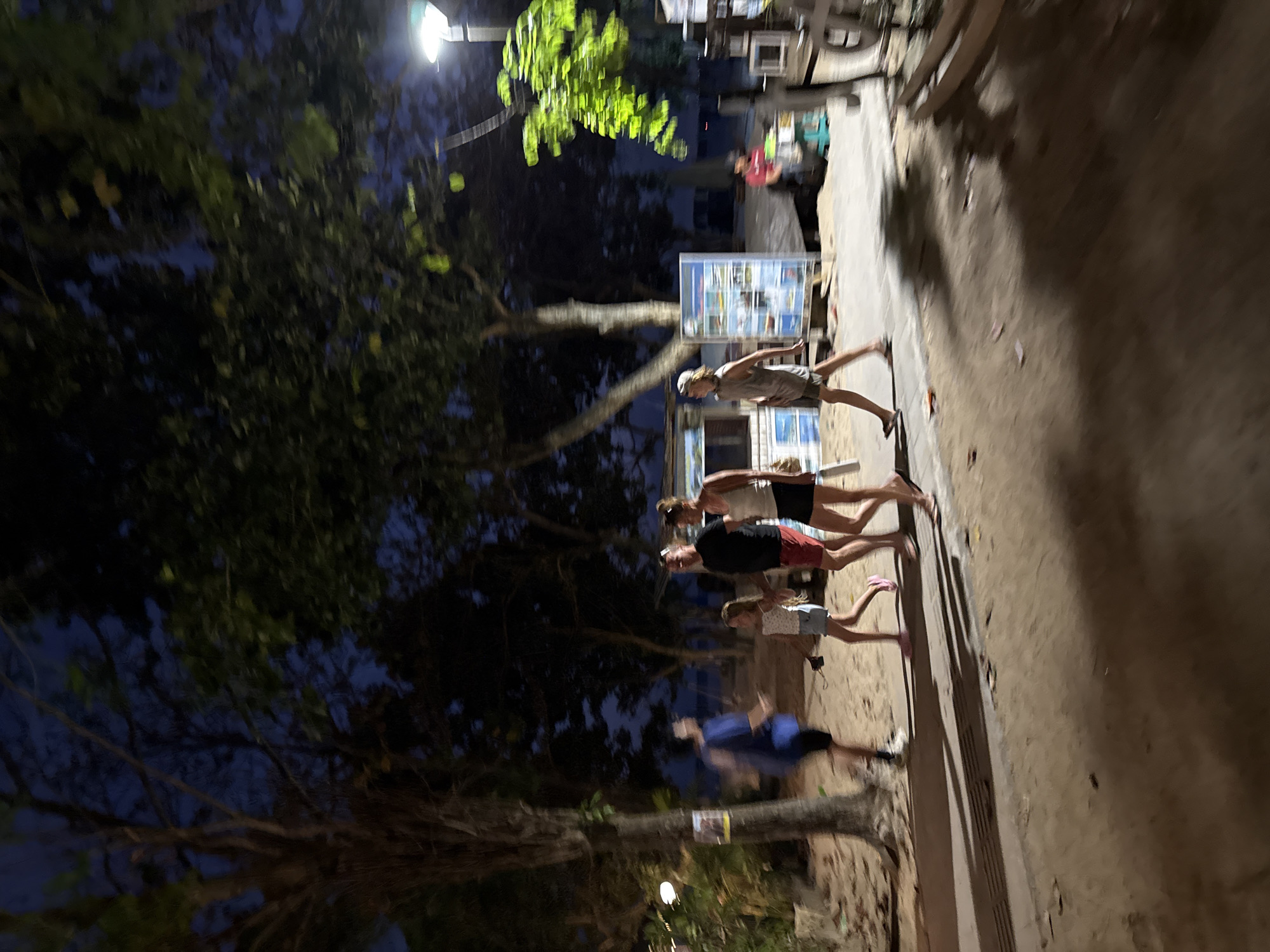 The image depicts a group of people walking along a sandy path at dusk in Denpasar, Indonesia. A bright overhead light casts long shadows on the ground, suggesting it is nighttime or very late evening. Trees with lush green foliage are visible on the left side of the frame, some of which are illuminated by the artificial light. In the background, there are structures that appear to be shops or stalls, with some signage visible. The people are casually dressed, wearing shorts and t-shirts, indicating a warm climate. Their postures suggest they are walking or standing, and their faces are not clearly visible due to the low light and motion blur. The overall atmosphere is casual and relaxed, typical of a beachside or tourist area at the end of the day. There are no discernible emotions captured due to the image quality. The ground is a sandy, earthy texture, with scattered debris and fallen leaves.