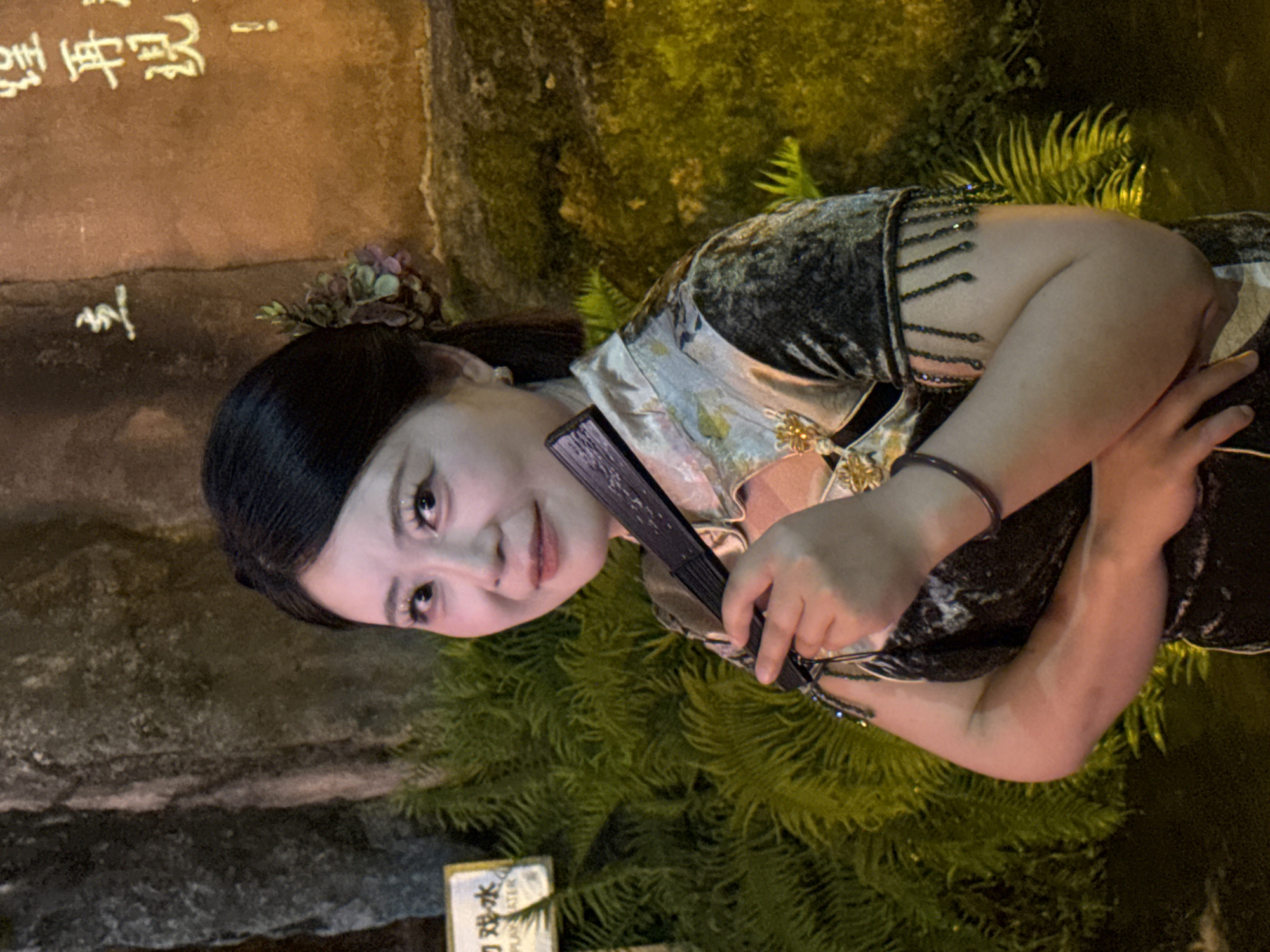 A medium shot captures a woman of East Asian descent, positioned in the foreground against a backdrop of a textured rock wall with Chinese calligraphy and lush green foliage. The woman is facing slightly towards the viewer, her head tilted. She has dark hair pulled back, adorned with a floral hairpiece on her right side. Her makeup includes bright eyeshadow and eyeliner. She is holding a black folding fan in her right hand, bringing it close to her face. She is wearing a traditional Chinese dress, a qipao, with a high collar and intricate patterns. The dress is dark with lighter, embroidered details around the neckline and sleeve cuffs, which are embellished with beaded fringe. A dark bracelet is visible on her left wrist. The environment appears to be outdoors, possibly at night, given the lighting which casts a warm glow on the rock and foliage. To the left of the woman, a sign with Chinese characters and "PLAY ATER" is partially visible, suggesting a public area. The overall scene has a serene and elegant aesthetic.