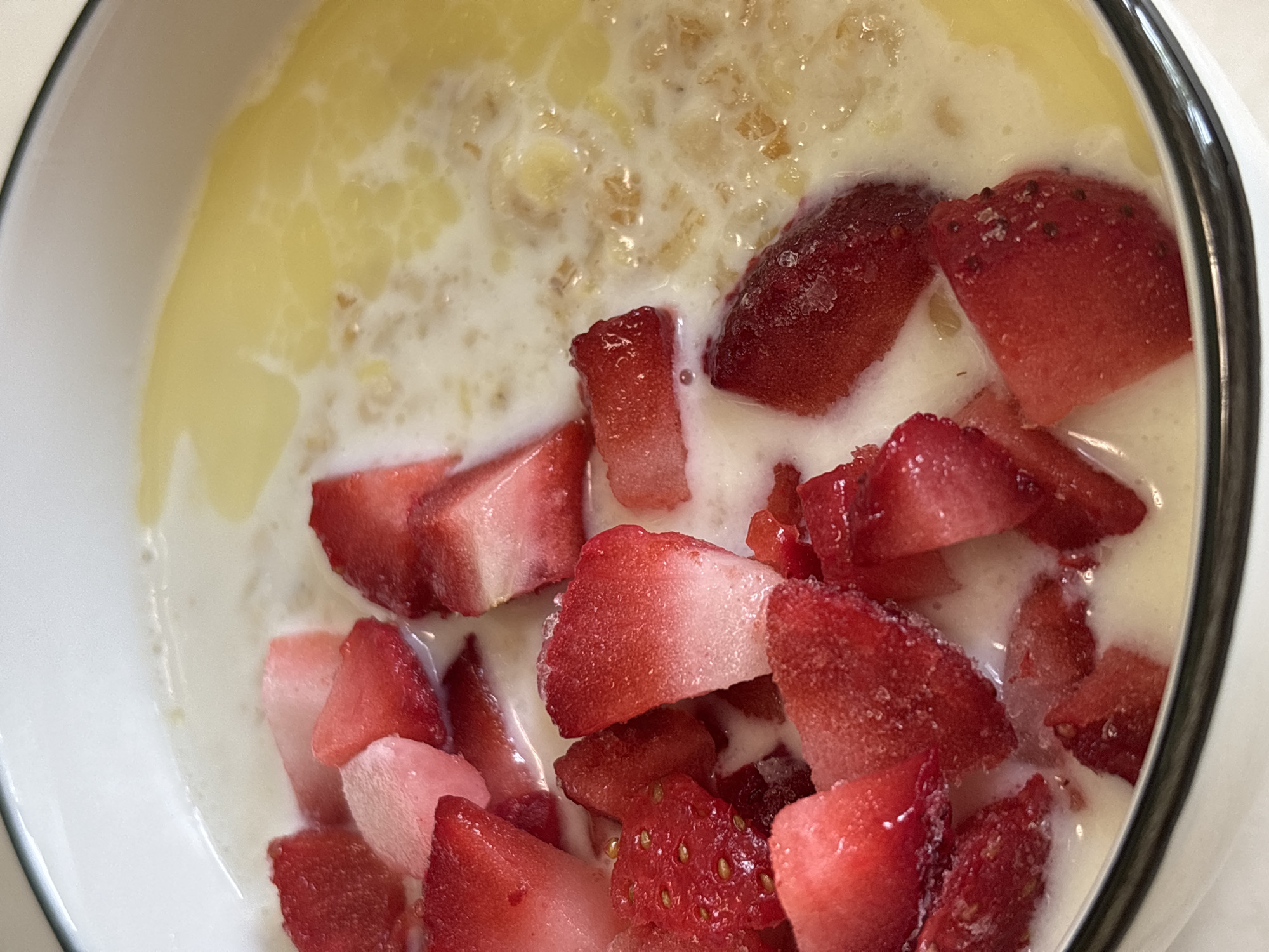 A high-angle, close-up shot depicts a bowl of oatmeal with sliced strawberries and a liquid, possibly milk or cream. The oatmeal appears to have a slightly yellow tint, possibly due to butter or another additive. The strawberries are predominantly red with some lighter pink tones. The bowl is white with a dark rim. The location of Rusanovo, Russia, is not discernible from the image.