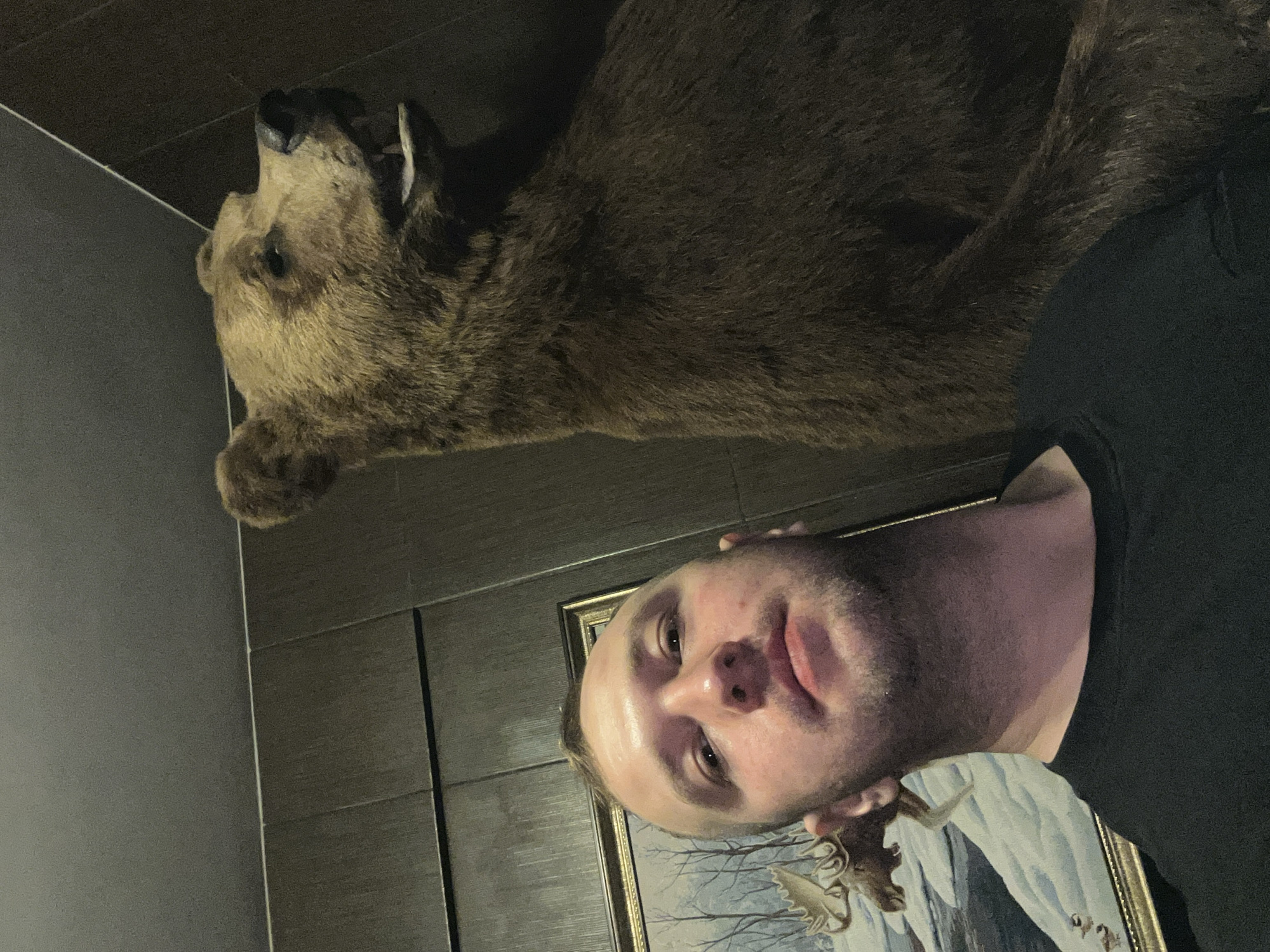 A man takes a selfie in front of a taxidermied bear head mounted on a wall in a room. The man has fair skin, short blonde hair, and a stubbly beard. He is wearing a black shirt. Behind him is a framed picture depicting a landscape with moose. The wall appears to be covered in dark tiles. The location is Chaikovsky, Russia.