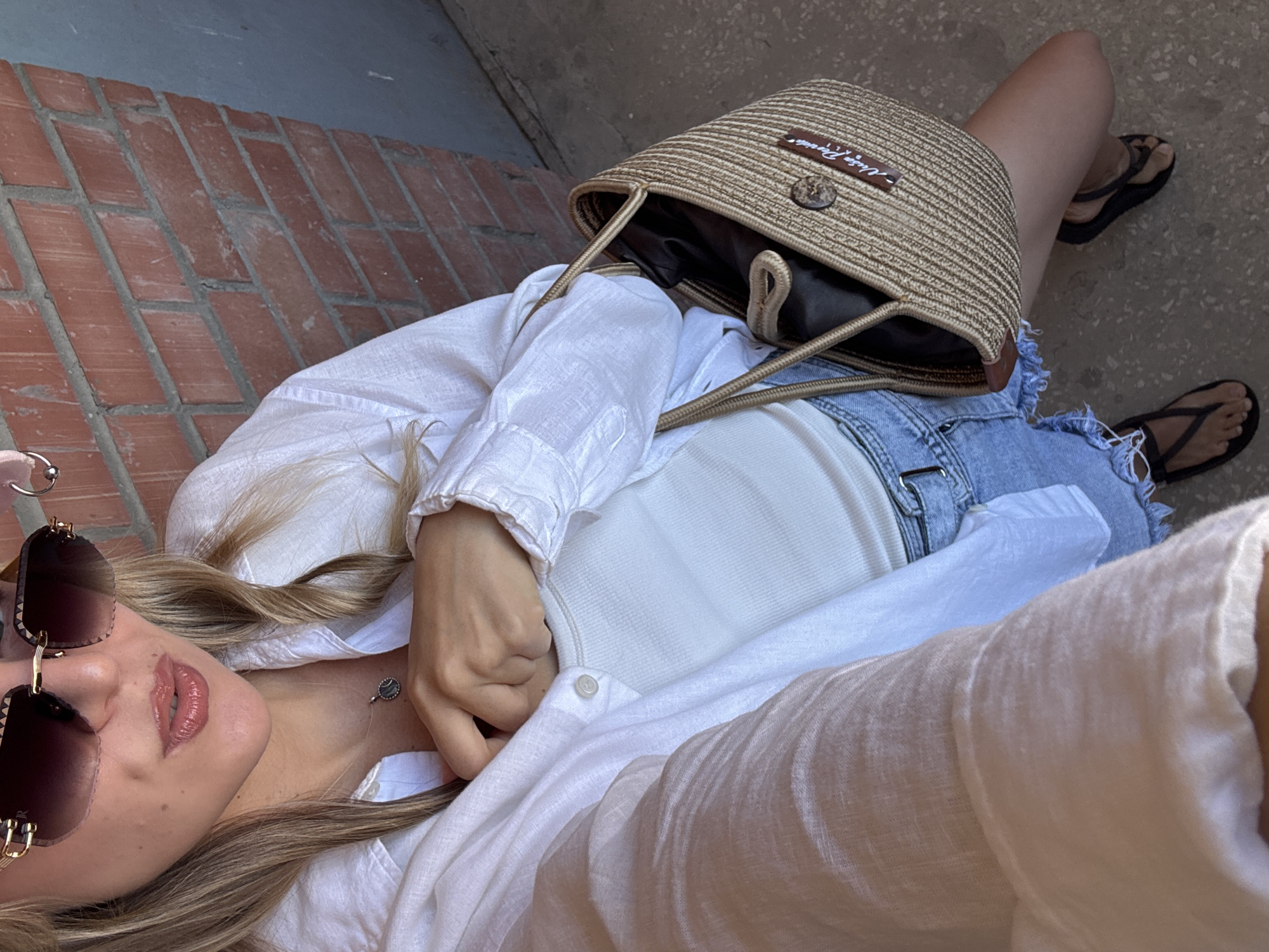 A young woman stands outdoors, leaning against a brick wall. She wears large sunglasses, a white linen button-down shirt over a white tank top, denim shorts, and dark flip-flops. A woven straw bag with leather straps is slung over her shoulder. Her left hand is tucked into the front of her shirt. The ground is a textured grey surface, and the background includes a brick wall and a grey wall. The lighting suggests it is daytime.