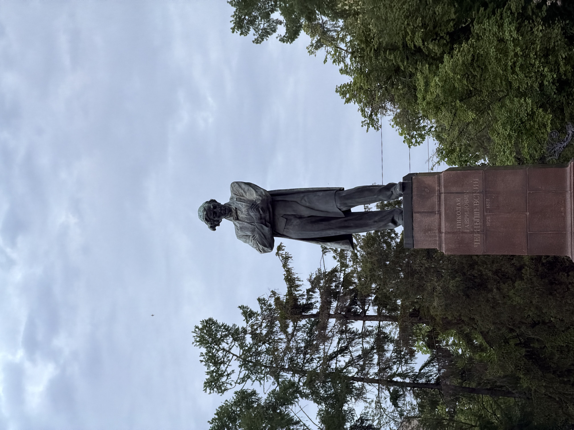 A low-angle shot depicts a statue of Nikolai Chernyshevsky in Saratov, Russia, against a cloudy sky. The statue stands on a brick pedestal inscribed in Russian with "Nikolai Gavrilovich Chernyshevsky 1828-1889". The statue is of a man in a long coat, with his arms crossed, and facing towards the left of the shot. The statue is surrounded by trees. In the sky, there is a small plane.