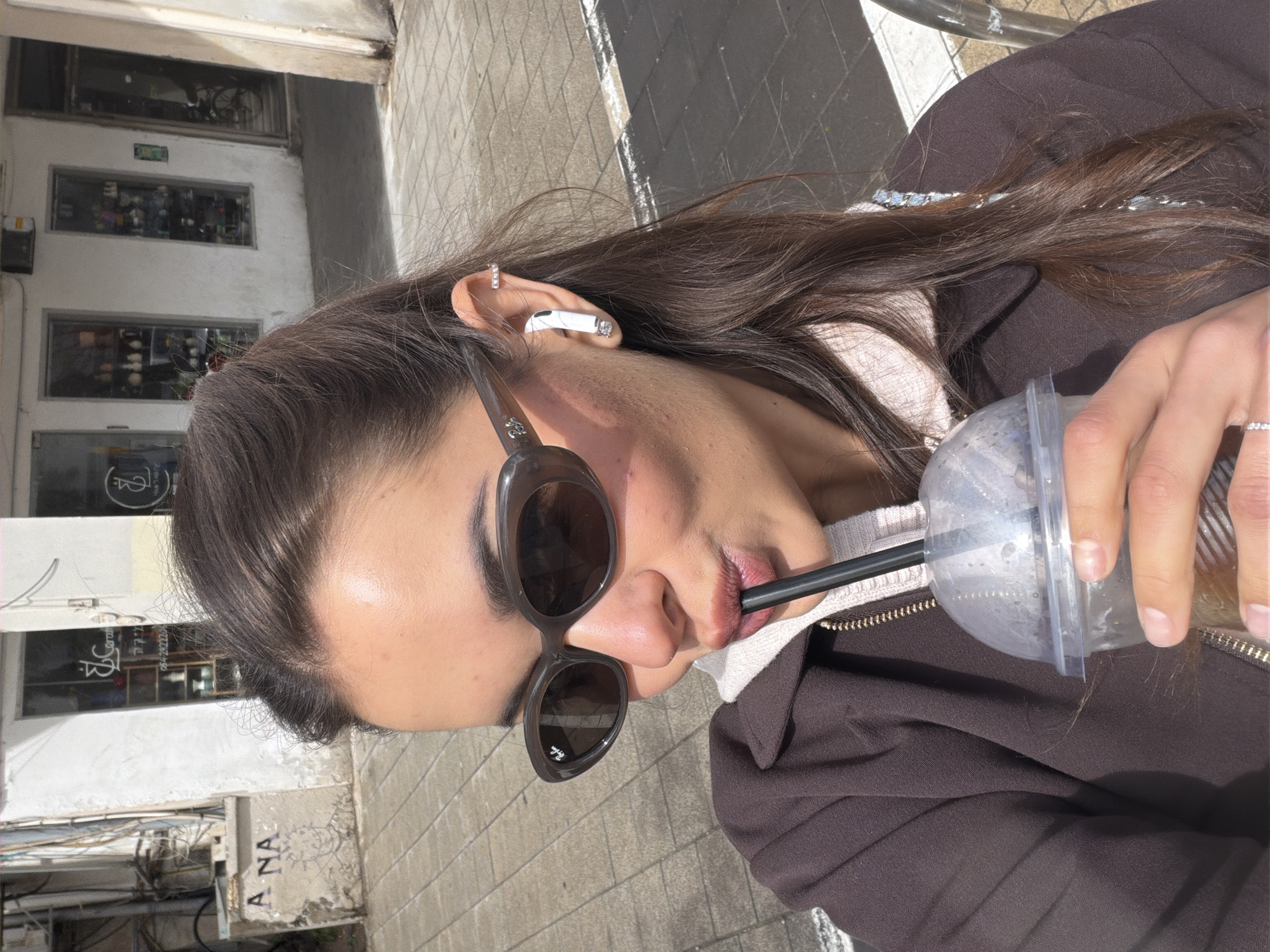 A female individual is depicted outdoors, positioned in the foreground and facing slightly towards the viewer. She has long, dark brown hair, partially secured, with loose strands visible. Her eyes are obscured by dark-tinted, oval-shaped sunglasses featuring a Ray-Ban logo on the left lens. In her right ear, she wears a white wireless earbud with a small, clear embellishment, alongside a minimalist silver-colored ear cuff on the upper cartilage. She is dressed in a dark brown collared jacket with a gold-toned zipper, worn over a lighter-toned hooded garment. Her right hand holds a clear plastic cup with a dome lid and a black straw, from which she is drinking, indicated by her pursed lips around the straw. The cup contains a brown liquid and shows condensation on its exterior. A silver-colored ring is visible on her right ring finger.
The background shows an urban environment with a light-colored paved sidewalk and a building constructed of light-colored stucco or concrete. The building features multiple glass storefronts. One storefront on the left displays a stylized "JC" logo, partial Hebrew text, and the phone number "054-2922248". This phone number is associated with JC Ceramics, located at Shlomo Hamelech 11, Tel Aviv-Yafo, Israel. Graffiti, including the letters "A NA," is visible on a concrete barrier or wall on the far left. A dark-colored bicycle is partially visible within an open doorway on the far right. The scene is illuminated by natural daylight.