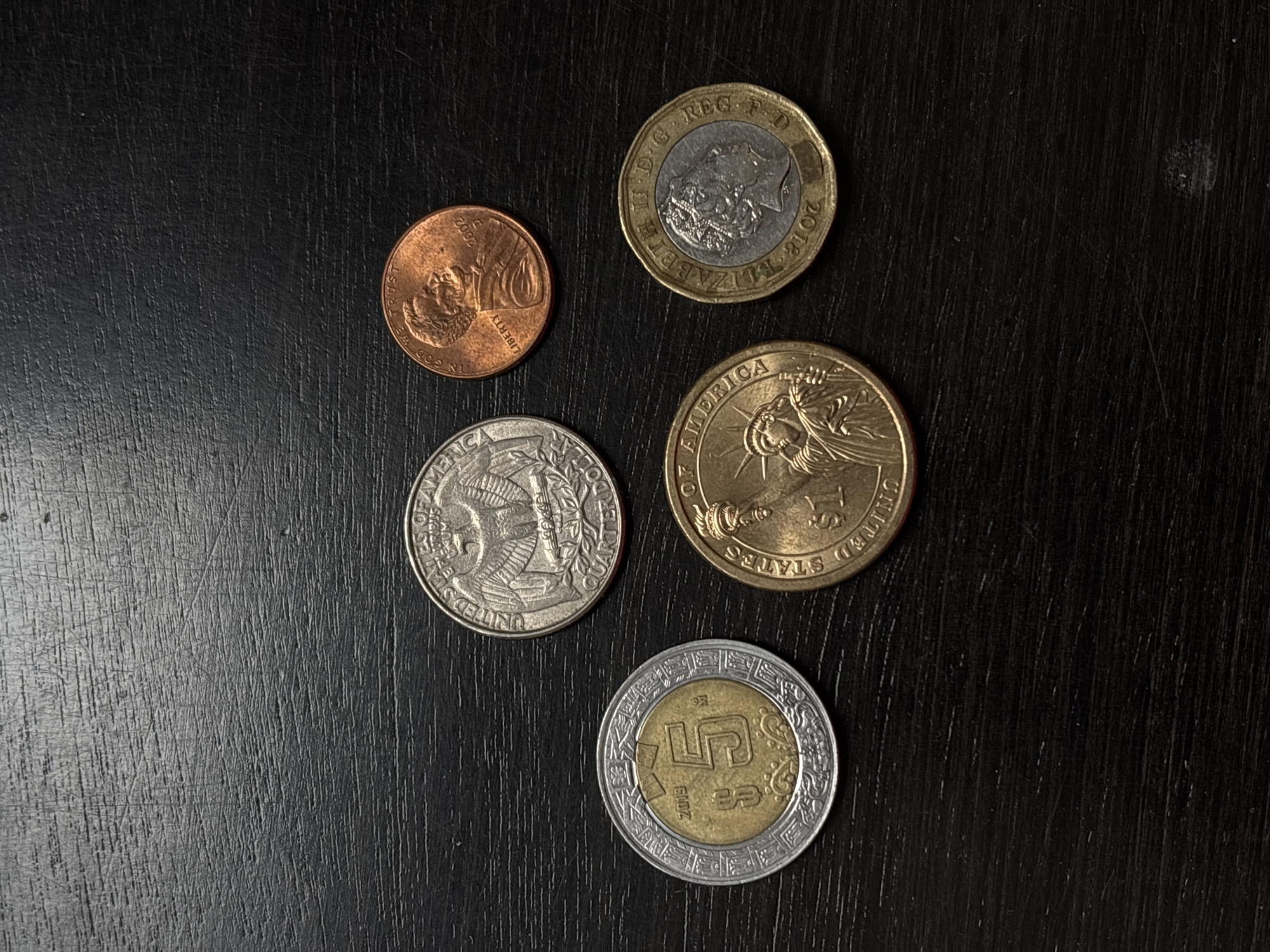 The image shows five coins arranged on a dark, textured surface. The coins, from top to bottom and left to right, include a United States penny, a British pound, a United States quarter, a United States dollar coin, and a Mexican 5-peso coin. The 5-peso coin indicates the location of Santa Clara Ocoyucan, Mexico.
