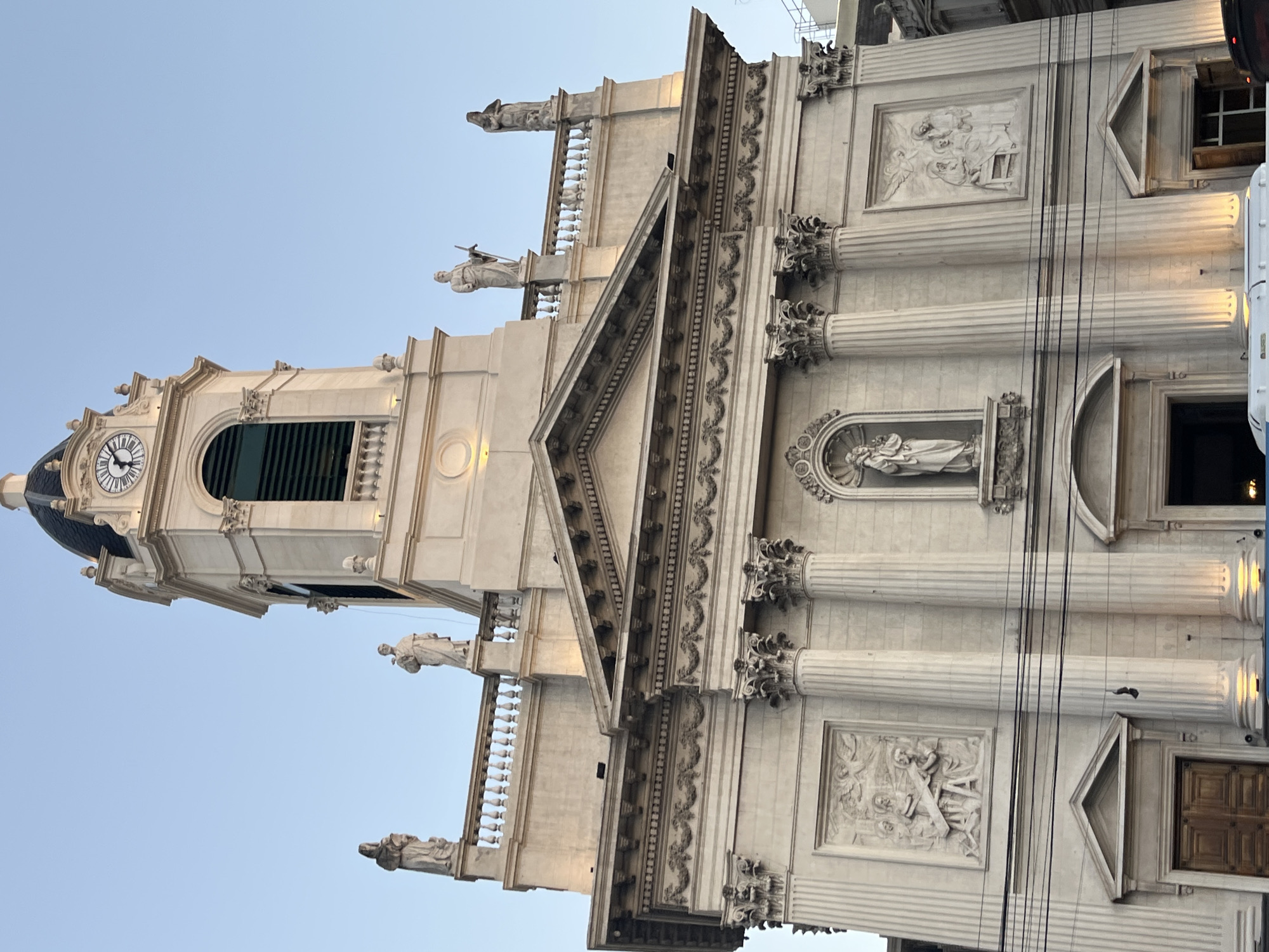 The image showcases a tall, ornate building with a clock tower, likely a church or cathedral, against a clear blue sky. The architecture is classical, featuring columns, statues, and decorative relief panels. The location is the Metropolitan Cathedral of Buenos Aires, Argentina. The image focuses on the exterior facade and does not depict any people. Wires are visibly obscuring parts of the building. The building is made of white stone.