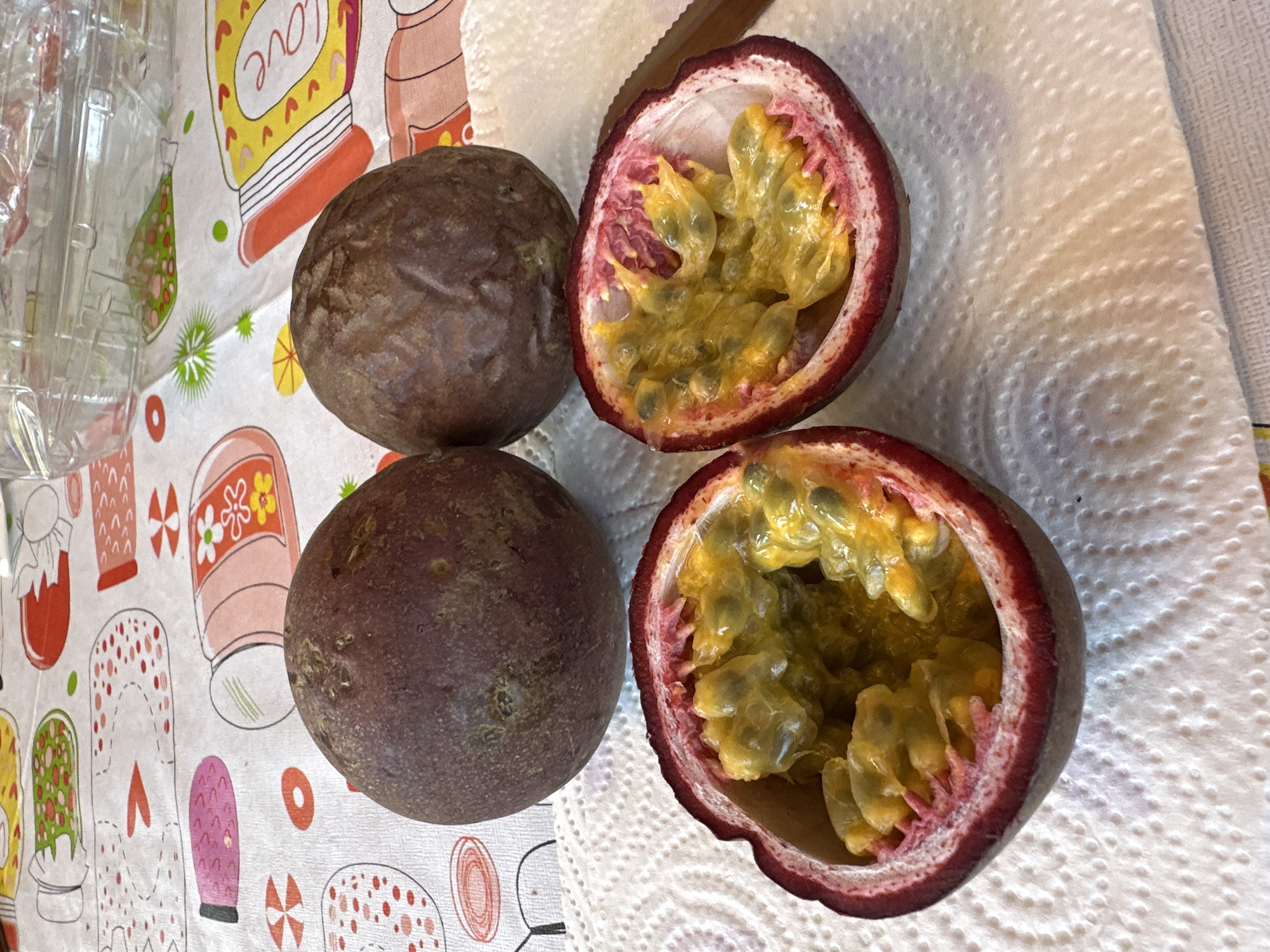 A close-up shot shows two whole passion fruits and two halves of a passion fruit on a white paper napkin. The passion fruits are dark purple and wrinkled. The cut halves reveal the yellow, pulpy interior with black seeds. The background features a colorful tablecloth with drawings of jars and fruit. The scene is indoors, on a table, with no discernable city or geographical markers of Borno, Italy.