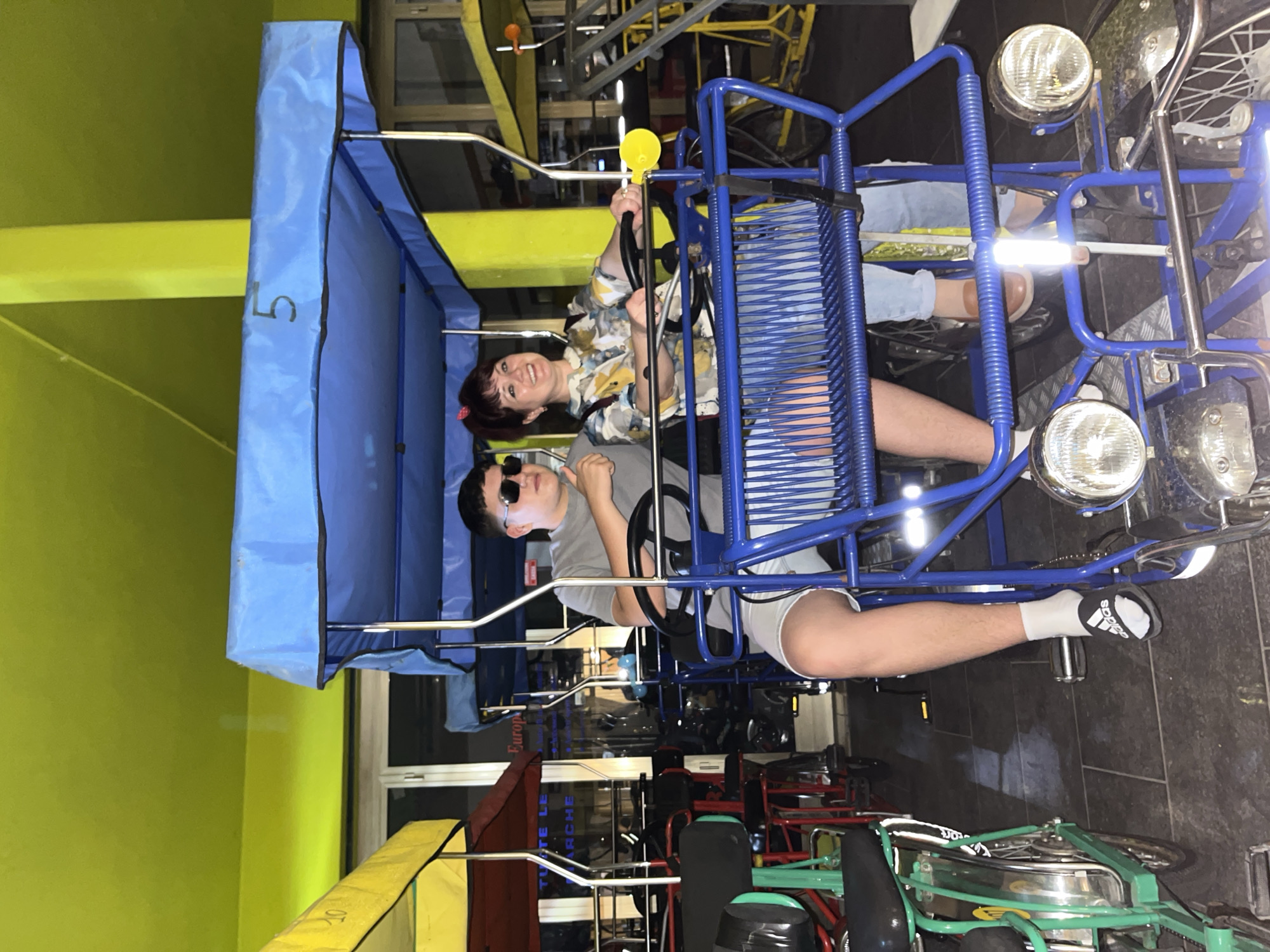 Two people are riding in a blue multi-person bike with a blue canopy marked with the number "5". The woman in the front is wearing a floral shirt and holding the steering wheel, while the person in the back is wearing a gray shirt and sunglasses. The multi-person bike has front headlights. Other similar bikes are visible in the background. The location appears to be a bike rental shop, potentially in the city of Jesolo, Italy. The environment includes a green wall.