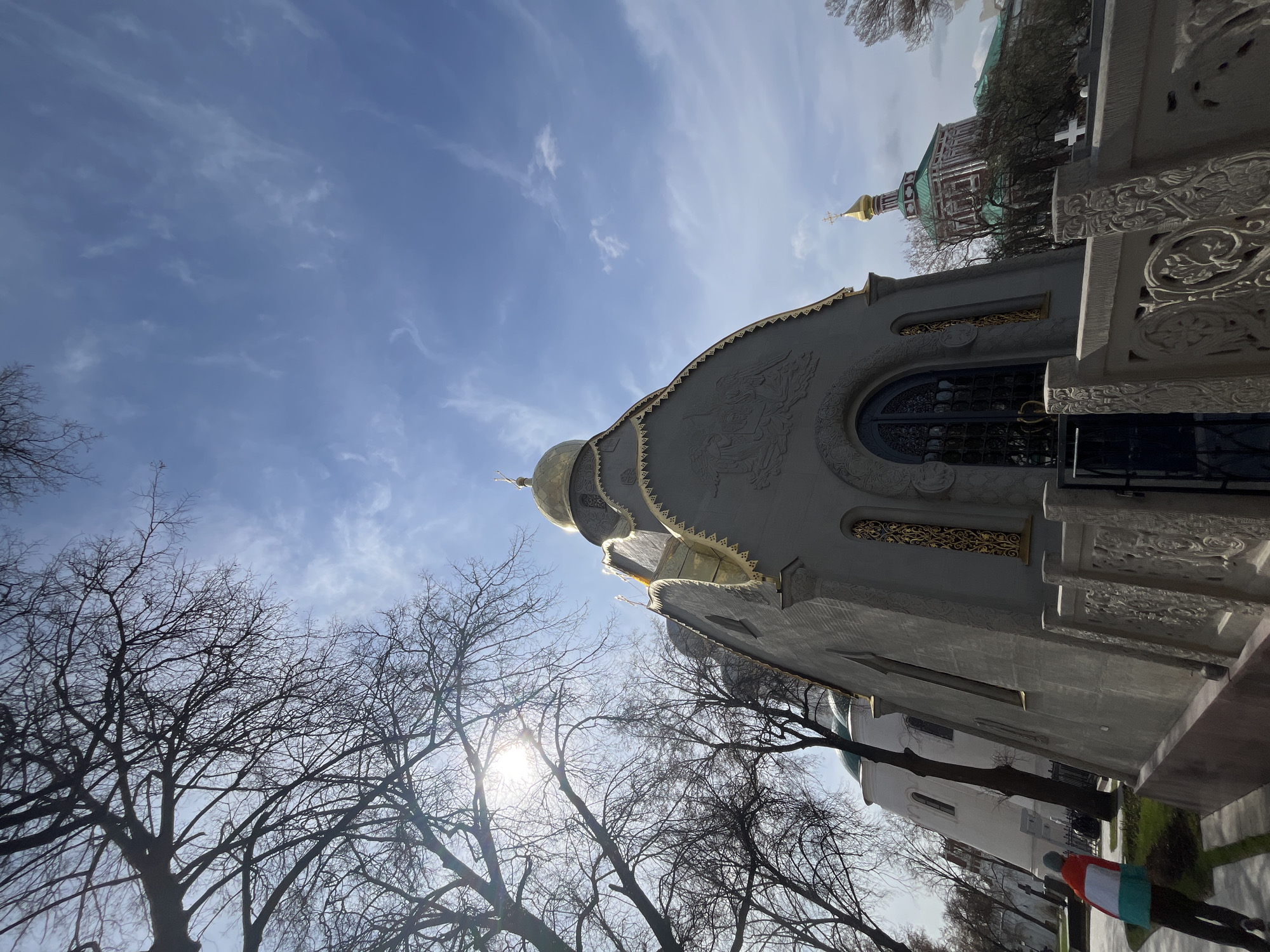 Captured from a low angle, the image shows a section of Moscow, Russia, with a distinct building featuring a rounded dome topped with gold. The dome is part of a gray structure decorated with intricate golden patterns. To the right, a wall showcases detailed carvings. In the background, a glimpse of a red and green structure is visible. Bare tree branches frame the lower part of the scene, obscuring a pedestrian with a white-red-green jacket and a glimpse of a white building on the lower right. The sky, a mix of blue and clouds, provides a backdrop. The sun is visible behind the trees.