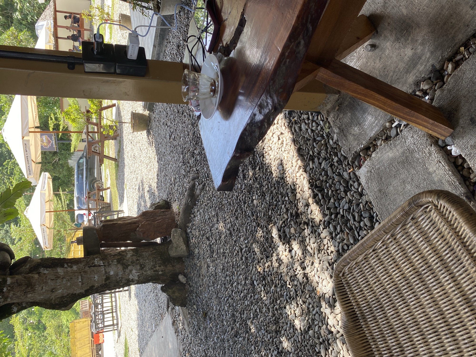 This image depicts an outdoor seating area at a coffee shop in Aluepiet, Indonesia. In the foreground, a woven chair is partially visible to the left, and a rustic wooden table with a live edge occupies the right side of the frame. On the table, an empty glass with remnants of a drink sits on a saucer, alongside a smartphone and charging cables plugged into an electrical outlet mounted on a yellow pillar. The ground is covered with small, gray pebbles, with a concrete path visible in the lower foreground. Behind the table, a large tree stands near a decorative stone arrangement. In the background, picnic tables, umbrellas, and a dark SUV are visible. Further in the distance, a building with people sitting inside and lush greenery suggest the broader environment of the establishment. The lighting indicates it is daytime.