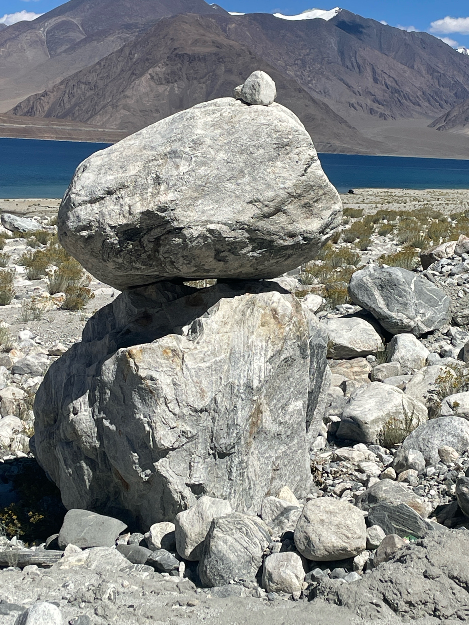 This is a full, eye-level shot of a rock cairn on a rocky shore with a body of water and mountains in the background. The cairn consists of three large, grey, unevenly shaped stones stacked on top of each other. The largest stone is at the bottom, followed by a slightly smaller stone, and a small, roundish stone on top. The rocks are rough and textured, with visible cracks and fissures.
The environment is arid, with sparse, dry vegetation scattered around the rocks. The ground is covered with smaller stones and pebbles. In the background, there is a large, turquoise body of water, likely a lake, stretching horizontally across the frame. Beyond the water, a range of imposing mountains rises, their slopes textured and colored in shades of brown and grey. The peaks of some of the mountains are capped with white snow. The sky is a bright blue with some scattered white clouds.
The image is taken in Man, India, a town located in the Zanskar valley of Ladakh, known for its stunning mountainous landscapes and high-altitude lakes. The scene suggests a natural, outdoor environment, possibly a tourist spot due to the presence of the cairn, which is often created by hikers or visitors. There are no people visible in the image. The lighting indicates it is daytime, with the sun casting shadows on the rocks.