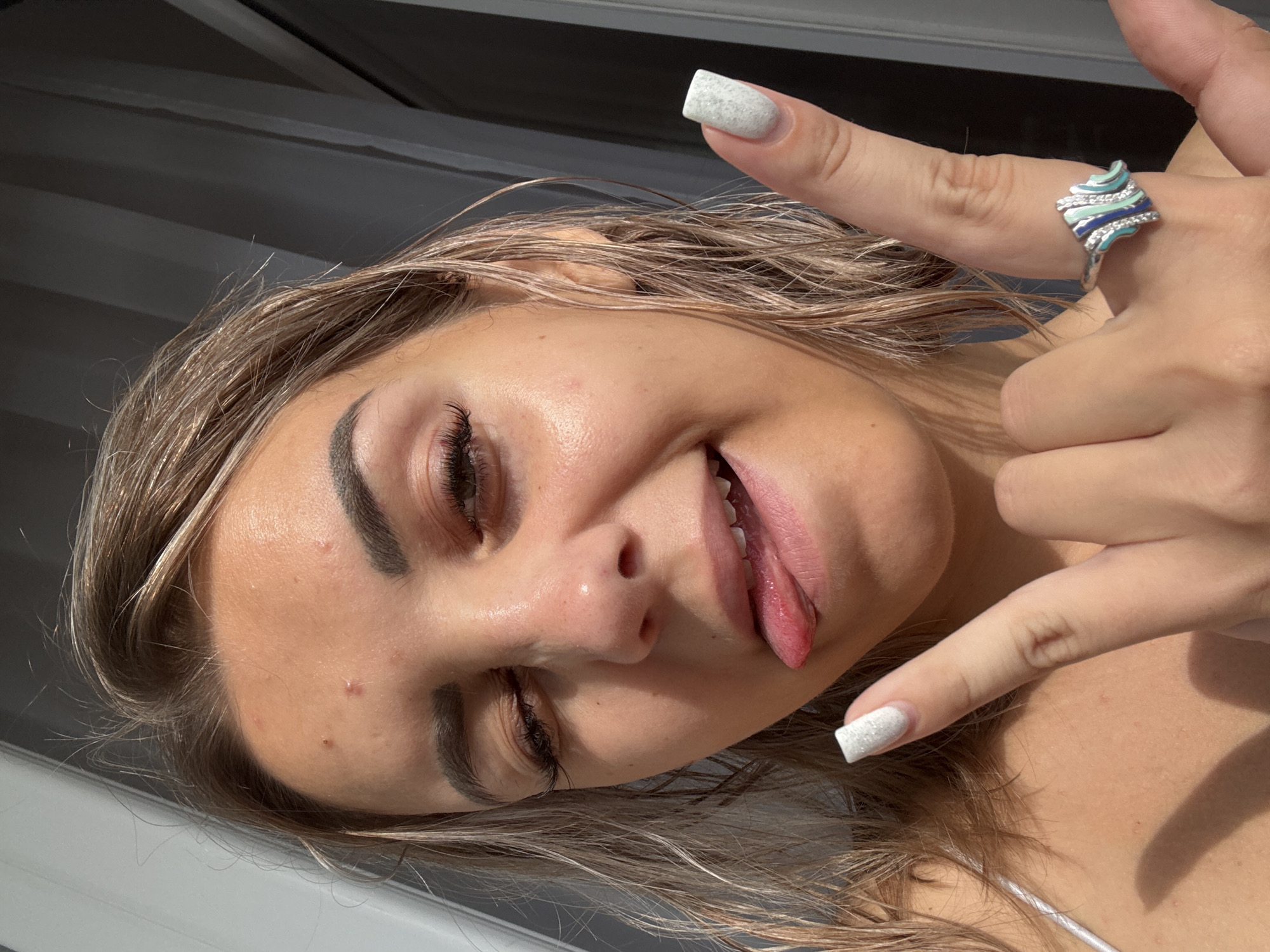 A young Caucasian woman is shown in a close-up, selfie-style shot. She has blonde hair, which appears wet and is styled loosely around her face. Her eyes are partially closed, and her tongue is sticking out. She is wearing a white top, and her left hand is raised, with her index and pinky fingers extended in a "rock on" gesture. She is wearing a ring on her left ring finger. There are several small blemishes on her forehead. The background appears to be indoors with natural light coming from the right, creating shadows on the left side of her face.