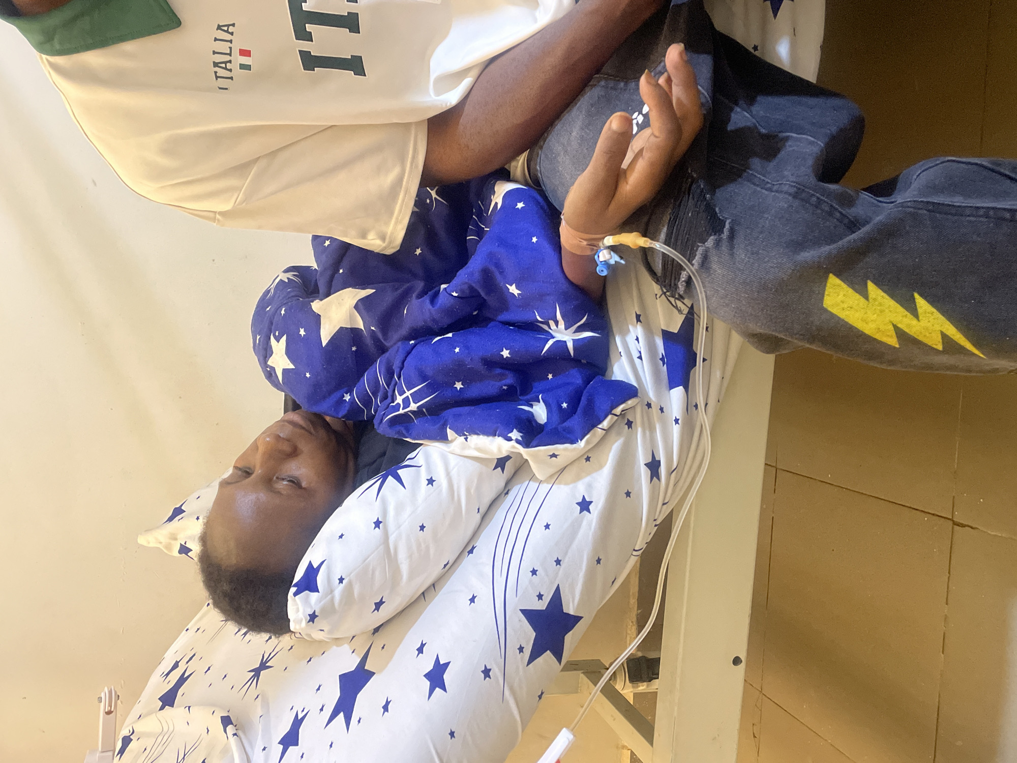 An individual is lying in a hospital bed. The bed is covered in white sheets with a pattern of blue stars. A blue and white comforter also with star patterns is partially covering the individual. An IV line is visible inserted into the individual's right arm. The person is wearing a light-colored shirt and dark jeans with a yellow lightning bolt design. Another individual is sitting next to the person in bed, with their arm visible. The setting is a hospital room, evidenced by the bed and IV equipment. The image was taken in the city of Gwong, Nigeria.