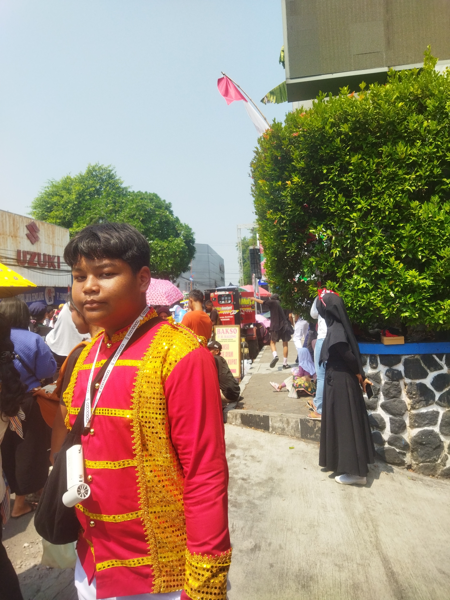 This is a medium shot photo taken outdoors on a sunny day. In the foreground, a young person, likely male, is standing and looking towards the camera. They are wearing a vibrant red and gold ceremonial outfit adorned with sequins and ornate trim, over a white shirt. A lanyard with a white fob is around their neck, and a black bag with a white personal fan is slung over their shoulder.
The background reveals a bustling street scene in Wonogiri, Indonesia. To the left, a faded "SUZUKI" sign is visible on a building, suggesting a commercial area. Further back, people are walking, some holding umbrellas. A food stall with the sign "BAKSO" (Indonesian meatball soup) and images of food is prominent.
On the right side of the image, a dense, green hedge lines a stone wall. Behind the hedge, a person in a black abaya and headscarf is visible, holding a dark object, likely a phone. Other individuals in casual attire are also present in the background, some sitting on the curb. The overall impression is of a lively event or street market in a city environment.