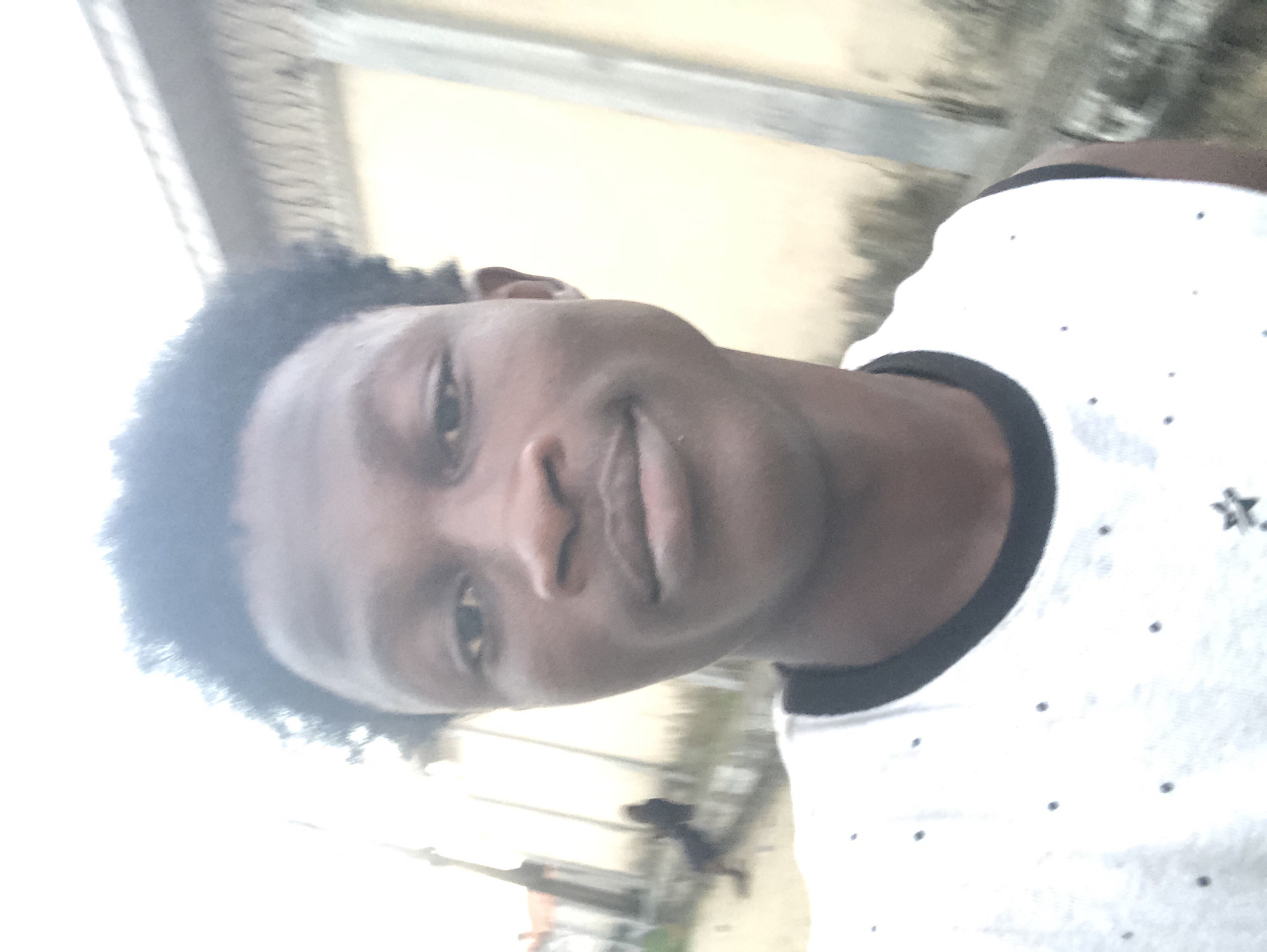 This is a close-up, selfie-style photograph of a young Black male wearing a white tank top with small dark dots. He has dark, short, curly hair and is looking directly at the camera with a slight smile. In the blurred background, a light-colored building with vertical lines can be seen, suggesting an outdoor urban setting. The overall lighting is bright, potentially indicating daytime. The background appears to be a street or pathway, with some greenery and structures visible in the distance, consistent with a location in Ado, Nigeria.