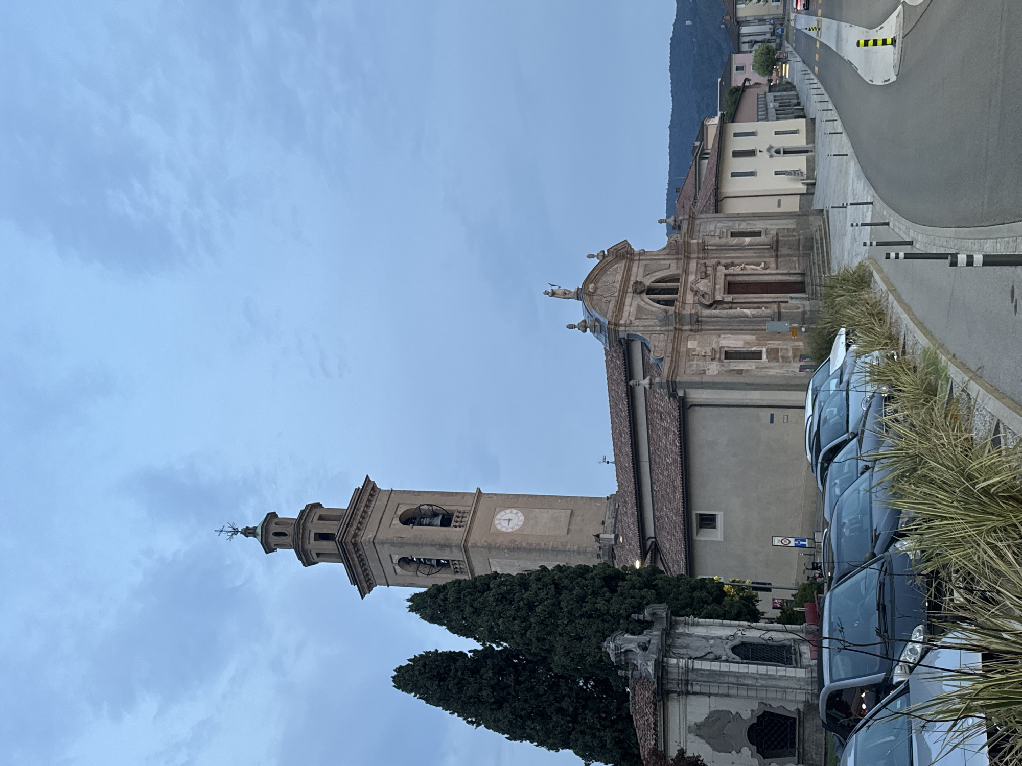 The image shows a view of the Church of San Lorenzo in Balerna, Switzerland, at dusk. A prominent bell tower with a clock face stands to the left of the church, which has a classical facade with a statue of a saint on top. Several tall cypress trees are situated to the left of the bell tower, and a decorative stone monument is also visible. In the foreground, a row of parked cars is lined up along a paved area, with some dry grass and foliage in front of them. A street curves away from the church and into the distance, with buildings lining its right side. The sky is overcast, with clouds suggesting the end of daylight.