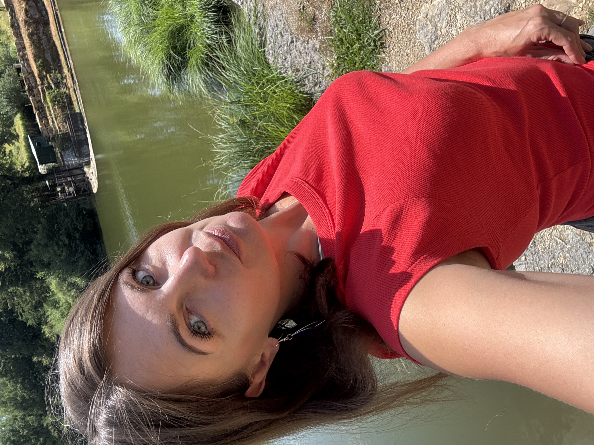 The media file is a portrait-style, horizontal, close-up photograph of a young woman lying down outdoors. She is wearing a bright red, ribbed short-sleeved top. Her head is tilted, and she is looking directly at the camera with a neutral expression. Her light blue eyes are a prominent feature, and she has long, brown hair that is spread out. One ear is visible, adorned with a silver earring. Her right arm is bent, with her hand resting on her chest, revealing a simple ring on her finger.
The background suggests a natural setting with a body of water to her left, likely a canal or river, with greenery and trees on the far bank. To her right, there is dry, rocky ground and more green grass. The lighting indicates it is daytime, likely sunny, casting shadows and highlighting the textures of her clothing and skin. The overall mood is relaxed and natural.
The location context provided is Coteaux du Lizon, France. There is no visible text in the image.