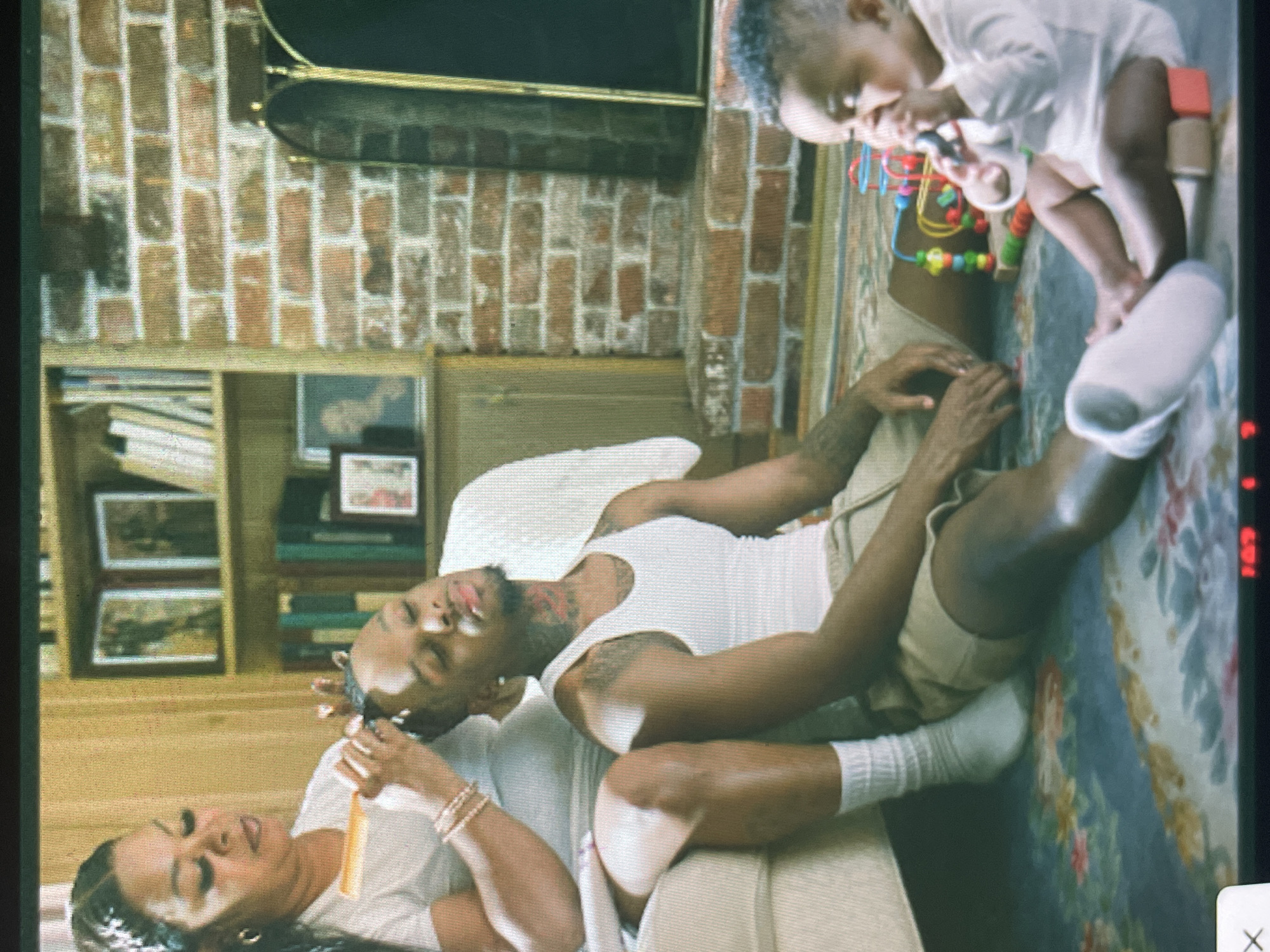 The image captures a warm, intimate domestic scene within what appears to be a living room in Sofia, Bulgaria. A man with a beard and tattoos on his neck and arms is lying in a relaxed posture, his head resting on a woman's lap. He is dressed in a white tank top, light brown shorts, and white socks. The woman, who has dark hair and multiple bracelets on her wrist, is wearing a white top and is gently combing the man's hair with a yellow comb.
To the right, a fair-skinned baby or young child, dressed in a white long-sleeved shirt and a diaper, is playing on a patterned rug. The child is engaged with various colorful toys, including a rattle, a bead maze, and wooden blocks.
In the background, a brick fireplace with a dark mantelpiece is visible. To the left of the fireplace, a wooden bookshelf holds several books and framed pictures, contributing to the cozy atmosphere. The room is well-lit, suggesting it is daytime. The overall impression is one of peaceful family connection and everyday life. No visible text is discernible in the image.
