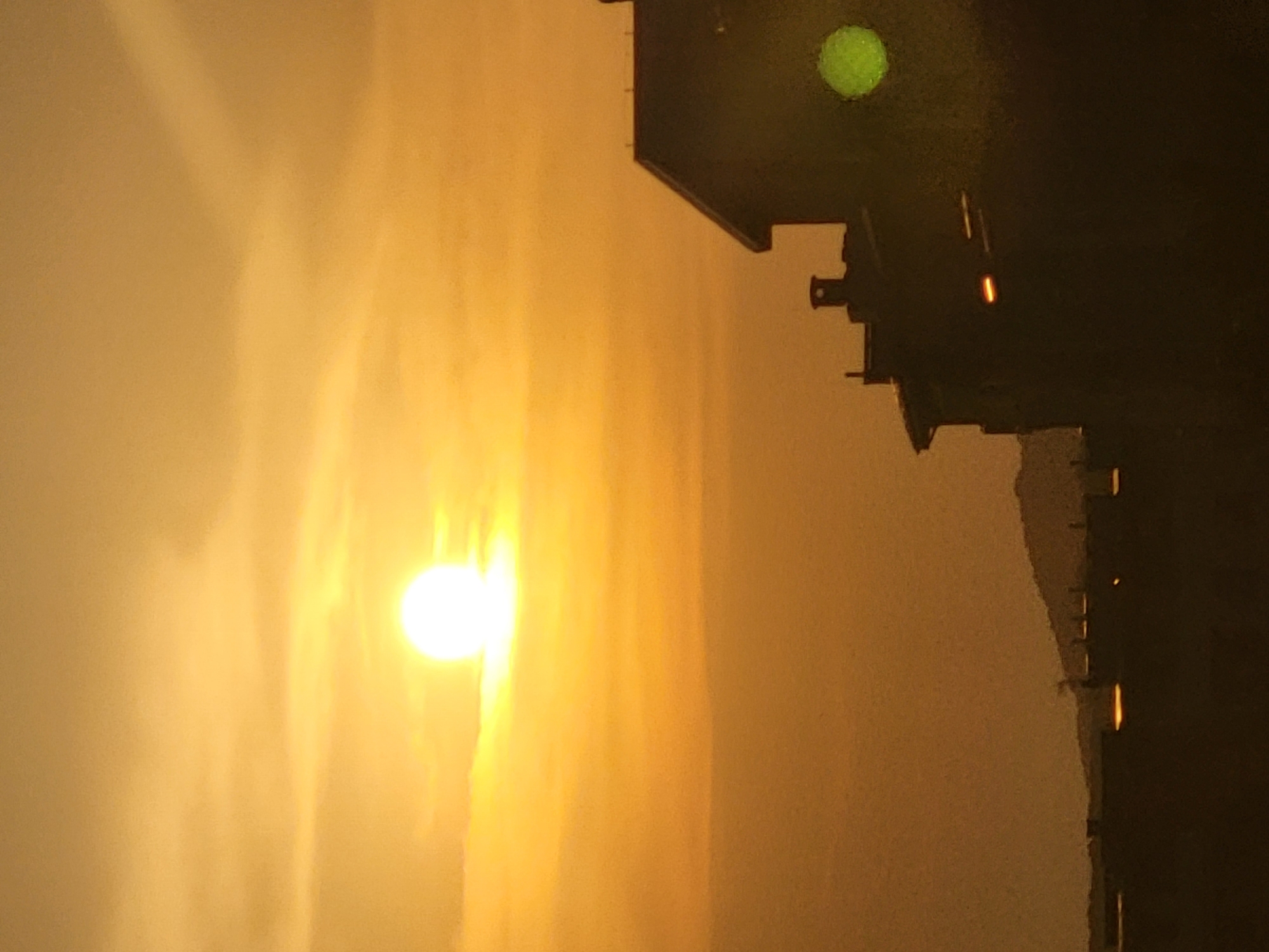 The image depicts a sunset or sunrise scene with a vibrant orange sky. The sun, a bright white orb, is visible on the left side of the frame, casting a warm glow. The right side of the image is dominated by a dark, silhouetted structure, possibly a building or a bridge, with several illuminated lights. One prominent light is a glowing orange bar, and another, higher up, appears as a faint green circle. The overall atmosphere is dramatic and perhaps a bit mysterious due to the contrast between the bright sky and the dark structure. The orientation of the image is vertical, suggesting it was taken with a phone in portrait mode or rotated later. There is no visible text in the image. The muted colors and soft focus of the sky suggest a hazy or cloudy atmosphere, common during twilight hours. The silhouette of the structure implies it might be a man-made object against the natural backdrop of the sky.