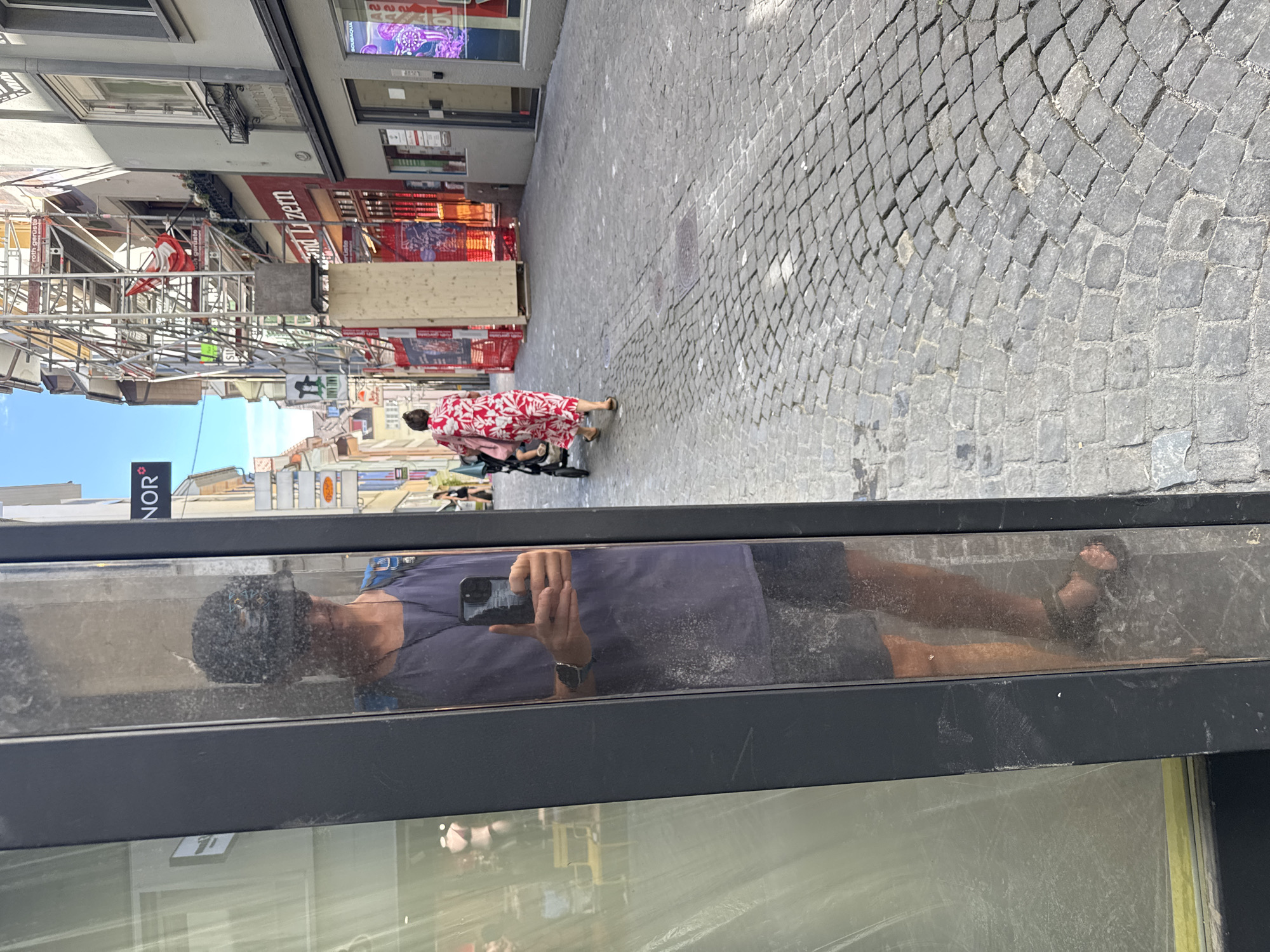 In the city of Lucerne, Switzerland, a person is seen reflected in a large glass storefront. The reflection shows the person wearing a dark baseball cap, sunglasses, a purple t-shirt, and dark shorts. They are holding a smartphone in their right hand, taking a selfie, and their left thumb is extended in a thumbs-up gesture. A backpack is visible over their left shoulder. The person is wearing a watch on their left wrist and dark sandals on their feet.
Beyond the reflection, the street is paved with cobblestones. A woman in a red and white floral dress is walking away from the viewer, pushing a stroller. In the background, buildings line the street. One building has scaffolding and what appears to be construction work in progress. Signs are visible on the buildings, including "NOR" and "land Luzern". The sky is blue and clear. The overall scene depicts a street in a European city on a sunny day.