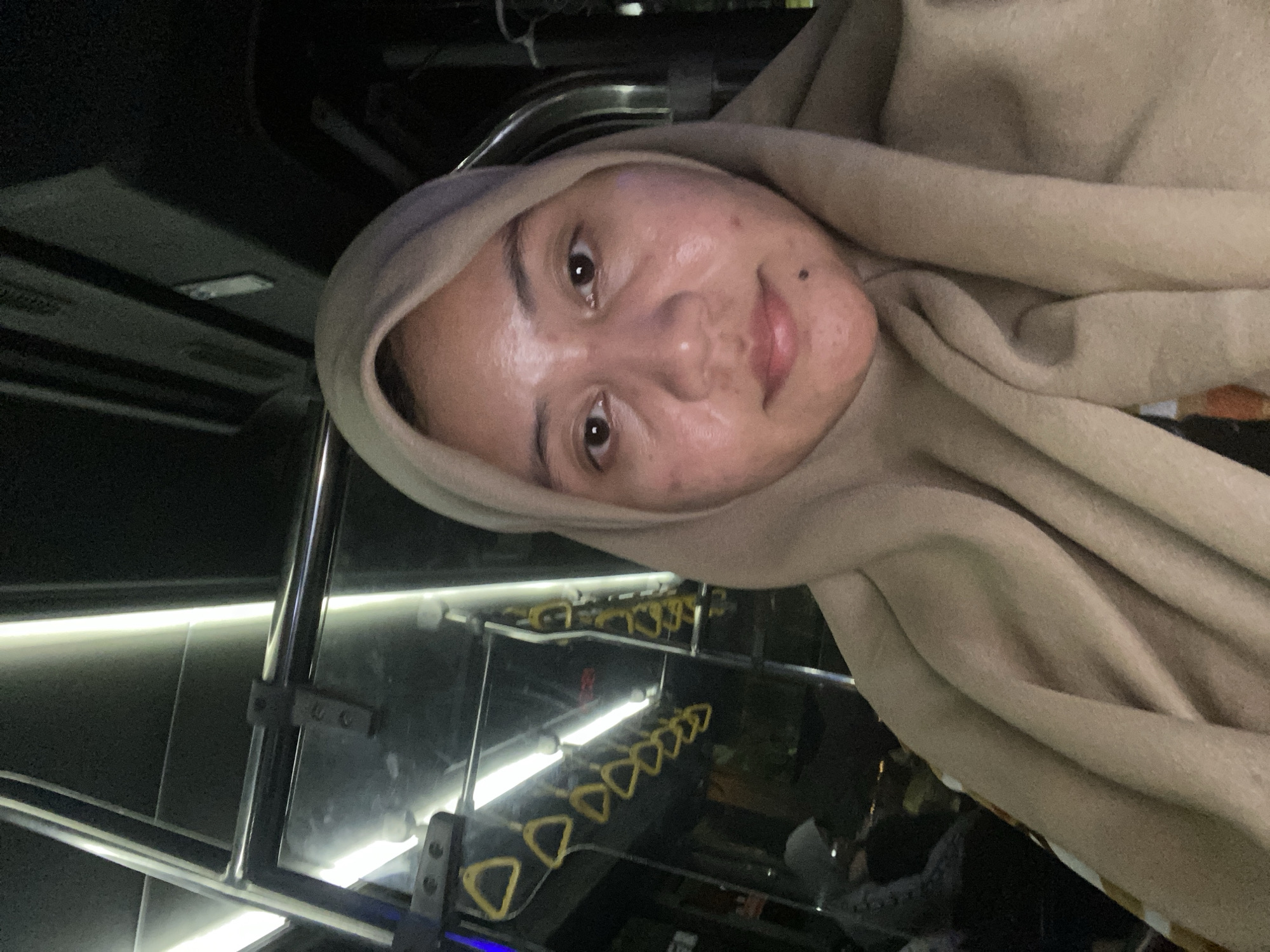 A woman wearing a beige hijab is taking a selfie inside a public bus at night. The bus appears to be traveling, as indicated by the motion blur in the background. She is seated, and the camera is angled upwards towards her face. The interior of the bus is visible, with fluorescent lights illuminating the ceiling and yellow grab handles hanging from a metal bar above. The woman's face is the primary subject, with her eyes looking directly at the camera. Her skin has some visible texture and blemishes. The background shows the dark interior of the bus, with other passengers blurred and indistinct. The lighting is dim, with the overhead lights creating bright streaks. The image is likely taken in Banjarmasin, Indonesia, given the context provided, but there are no specific landmarks visible to confirm the location.