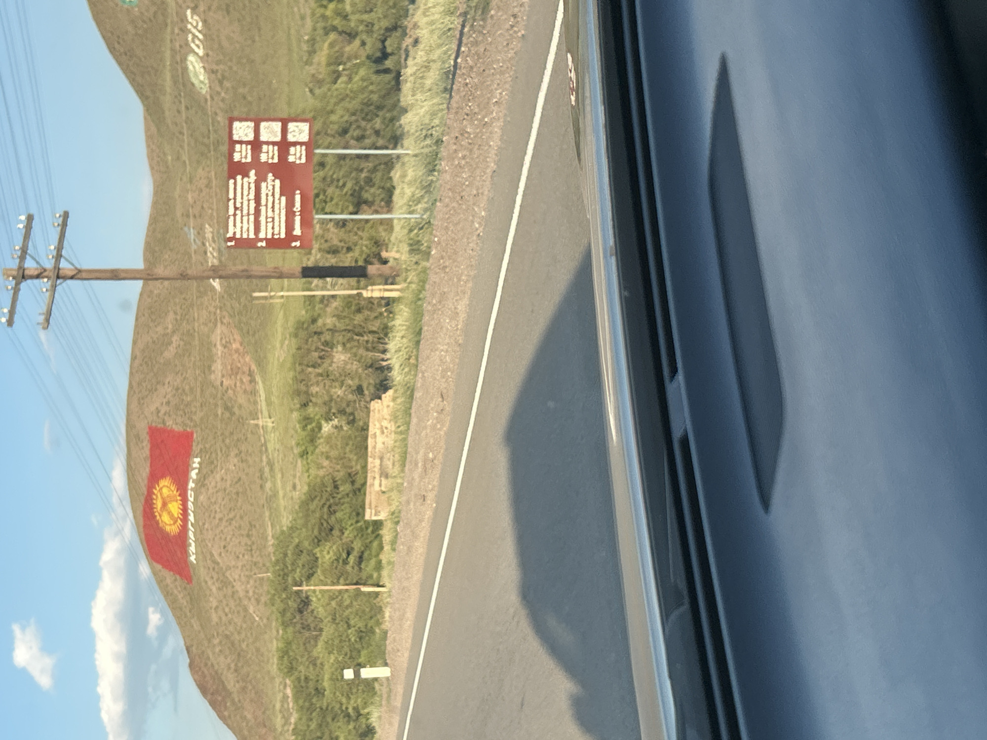 The image shows a scenic view of a hillside with the Kyrgyz flag painted on it, along with the word "КЫРГЫЗСТАН" in white letters below the flag. The location is identified as Kok Moynok Eki, Kyrgyzstan. A road runs in the foreground, and a shadow from a vehicle is visible on the road. There are trees and vegetation on the hillside. A wooden utility pole with wires stands prominently to the right of the flag. A red sign with text and QR codes is also visible near the utility pole. The sky is blue with some clouds, and the sun is shining, casting a shadow on the road. In the background, there are more hills. The image appears to have been taken from inside a vehicle, as indicated by the dashboard in the foreground.